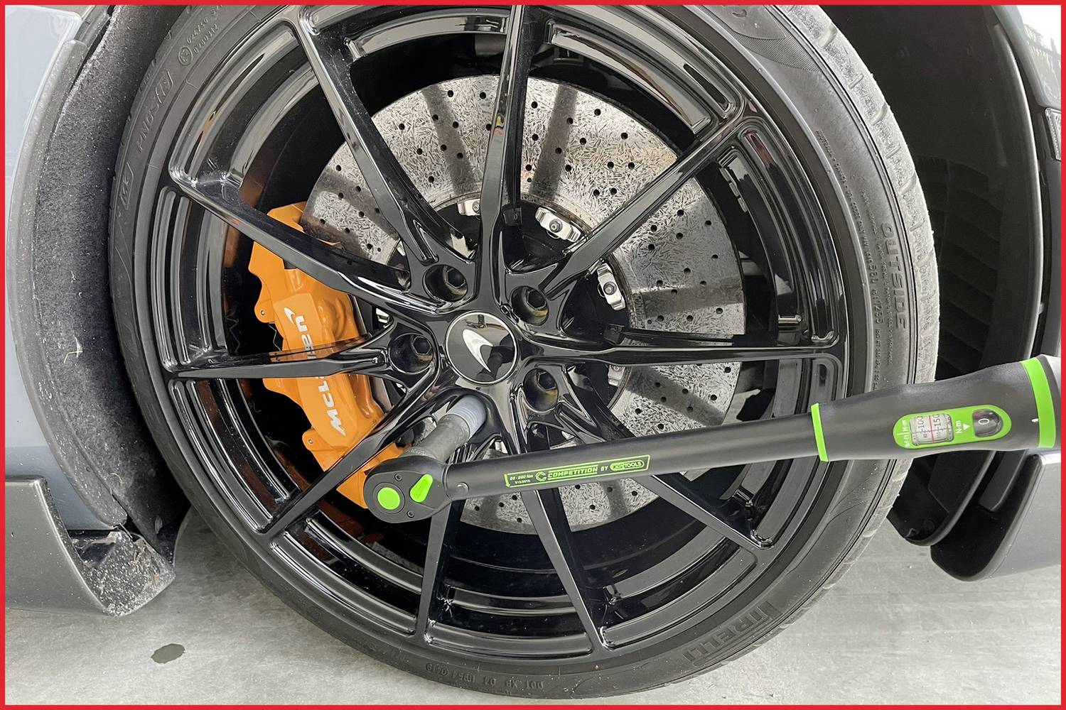 Schwarzes Autoreifen mit orangefarbenem Bremssattel und digitalem Drehmomentschlüssel, sichtbar auf Betonoberfläche. 