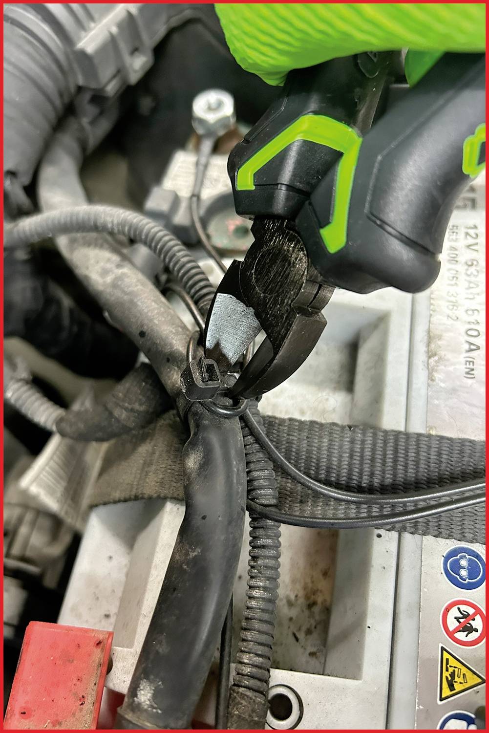 Eine Zange schneidet ein Kabel im Motorraum eines Fahrzeugs. Im Hintergrund sind Batterie und andere Motorbestandteile zu sehen.
