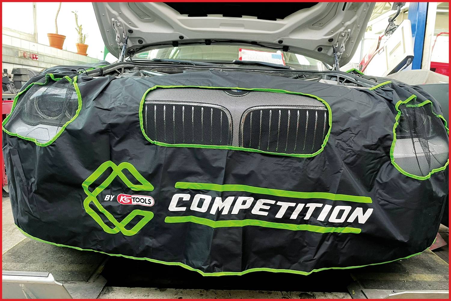 Auto mit geöffneter Motorhaube, abgedeckt mit 'COMPETITION' Werkstattschutzhülle.