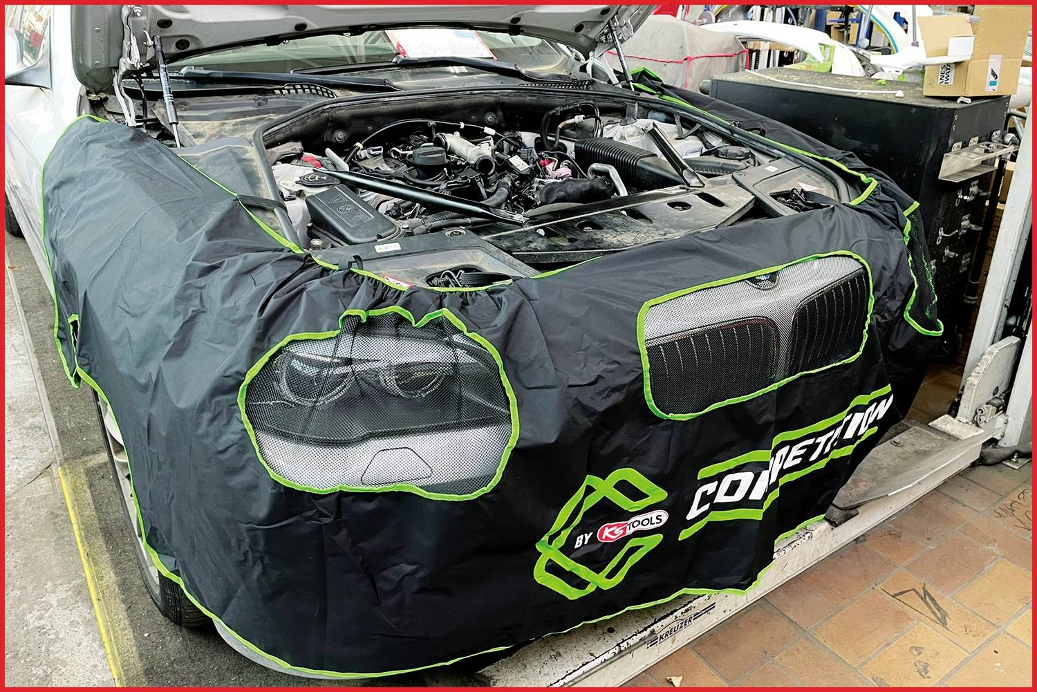 Auto in Werkstatt mit geöffneter Motorhaube; vordere Karosserie mit Schutzbezug abgedeckt. Wartungs- oder Reparaturarbeiten am Motor.