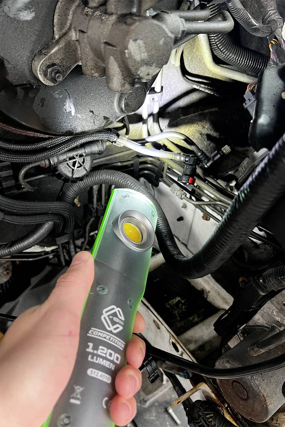 'Nahaufnahme einer Hand, die eine LED-Taschenlampe in einem Kfz-Motorraum hält. Verschiedene Kabel und mechanische Teile sind sichtbar.'
