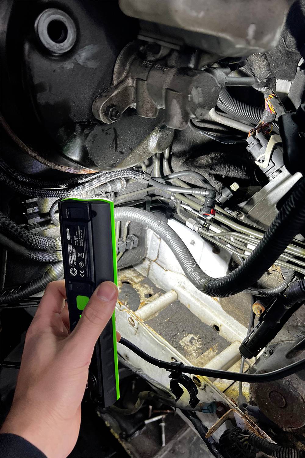 Eine Hand hält ein beleuchtetes Werkzeug unter der Motorhaube eines Fahrzeugs, um daran zu arbeiten oder zu inspizieren.