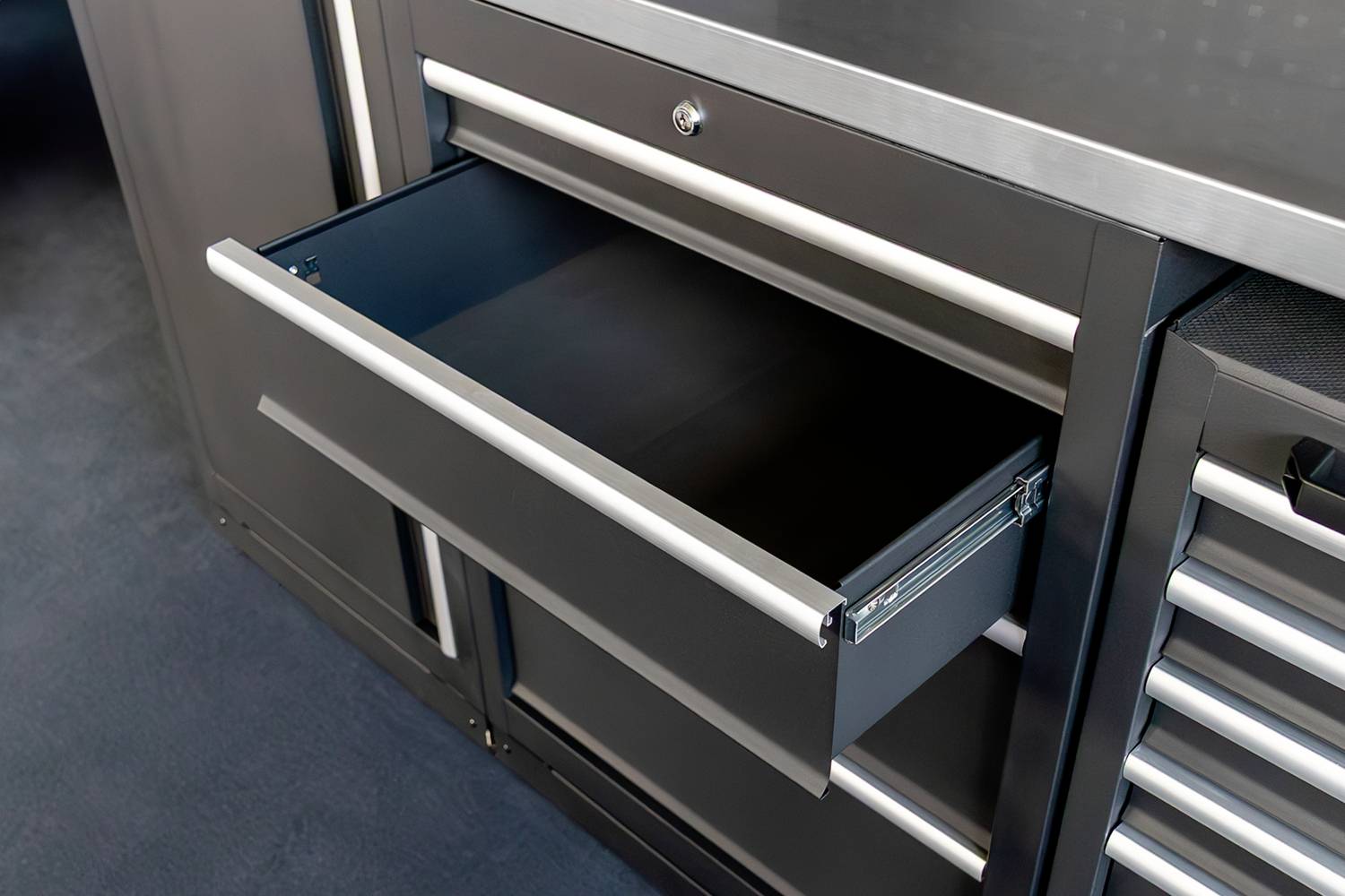 Eine geöffnete Schublade eines schwarzen Werkzeugschranks mit silbernen Griffen, vor einem Hintergrund aus Metall und Teppichboden.