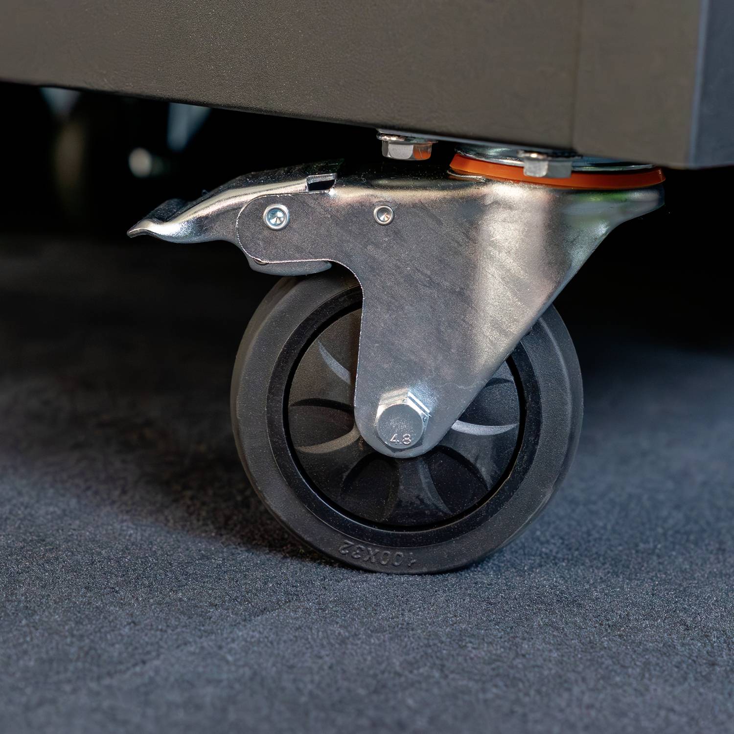 Ein graues Möbelrad auf Teppichboden, mit Bremse, an einem Möbelstück befestigt. Es zeigt eine robuste, industrielle Bauweise.