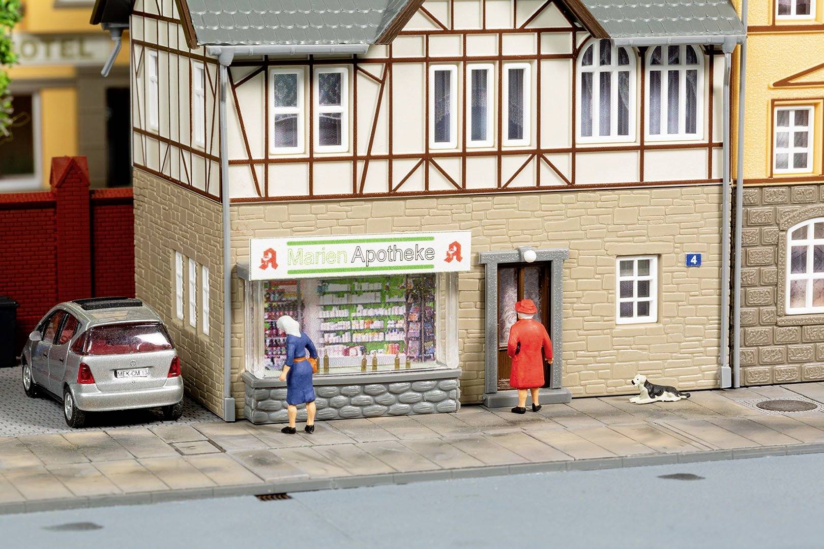 Zwei Personen stehen vor einem Schaufenster mit der Aufschrift 'Marien Apotheke'. Ein Hund liegt auf dem Gehweg daneben.