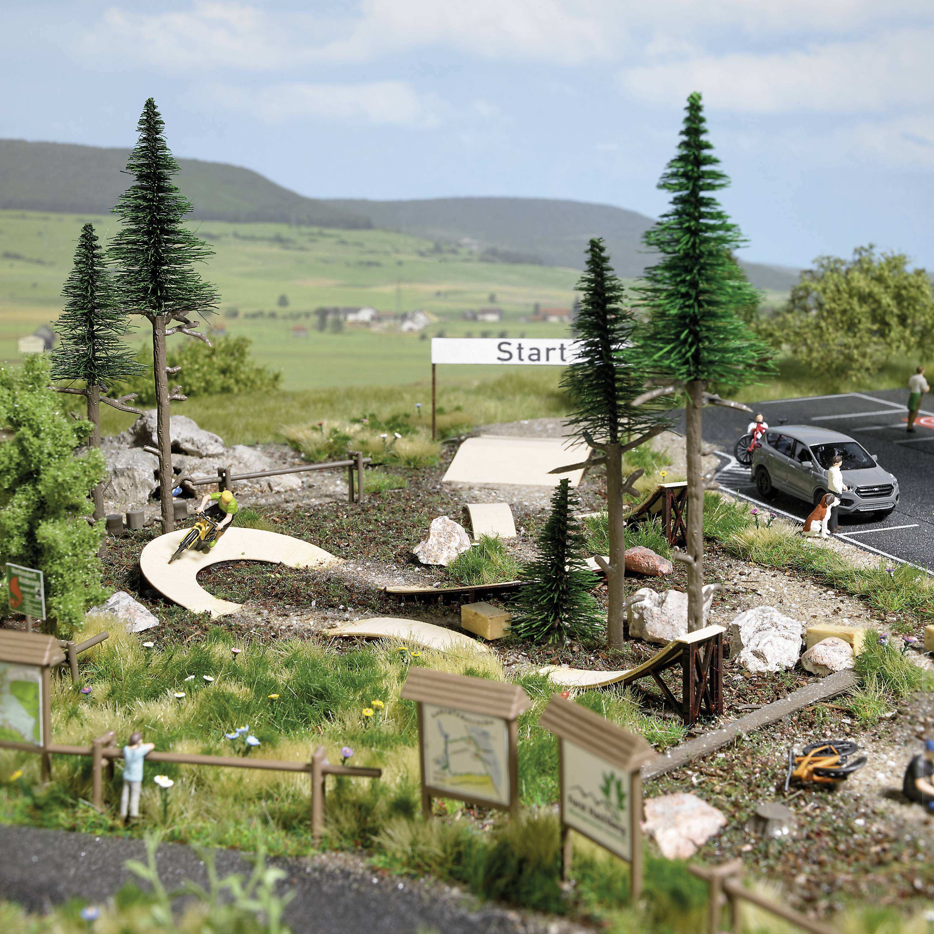Ein detailliertes Miniaturmodell zeigt einen Mountainbike-Park mit einem Fahrradfahrer auf einem kurvigen Weg, umgeben von Bäumen und Schildern, nahe einem Parkplatz mit Autos und Hügeln im Hintergrund.