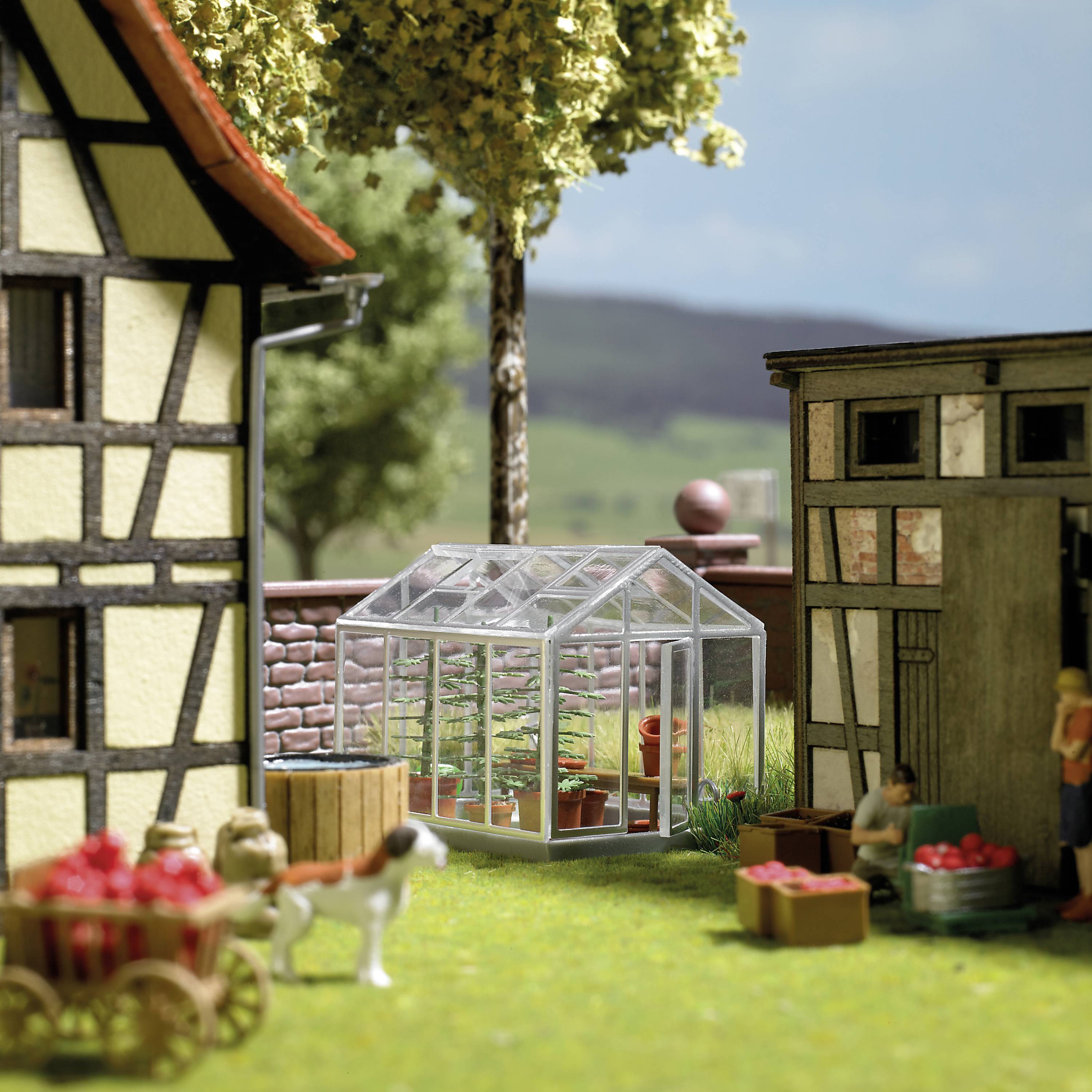 Miniaturmodell einer Dorfidylle mit Gewächshaus und Figuren, die draußen Äpfel ernten, vor ländlichem Hintergrund.