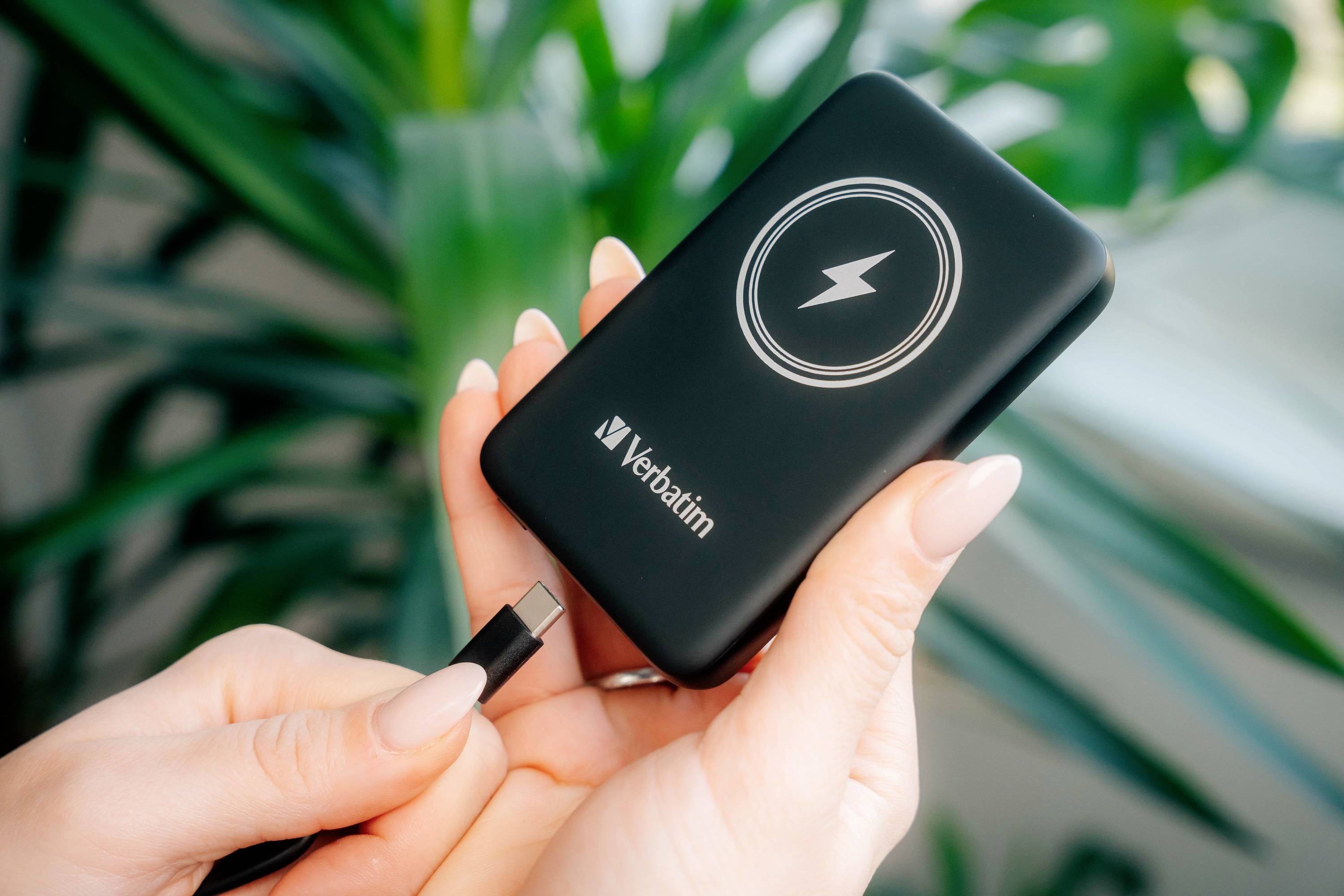 Eine Person hält eine tragbare Powerbank mit Blitzsymbol von Verbatim und steckt ein Kabel ein. Im Hintergrund sind grüne Pflanzen sichtbar.