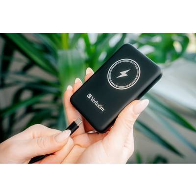 Eine Person hält eine tragbare Powerbank mit Blitzsymbol von Verbatim und steckt ein Kabel ein. Im Hintergrund sind grüne Pflanzen sichtbar.