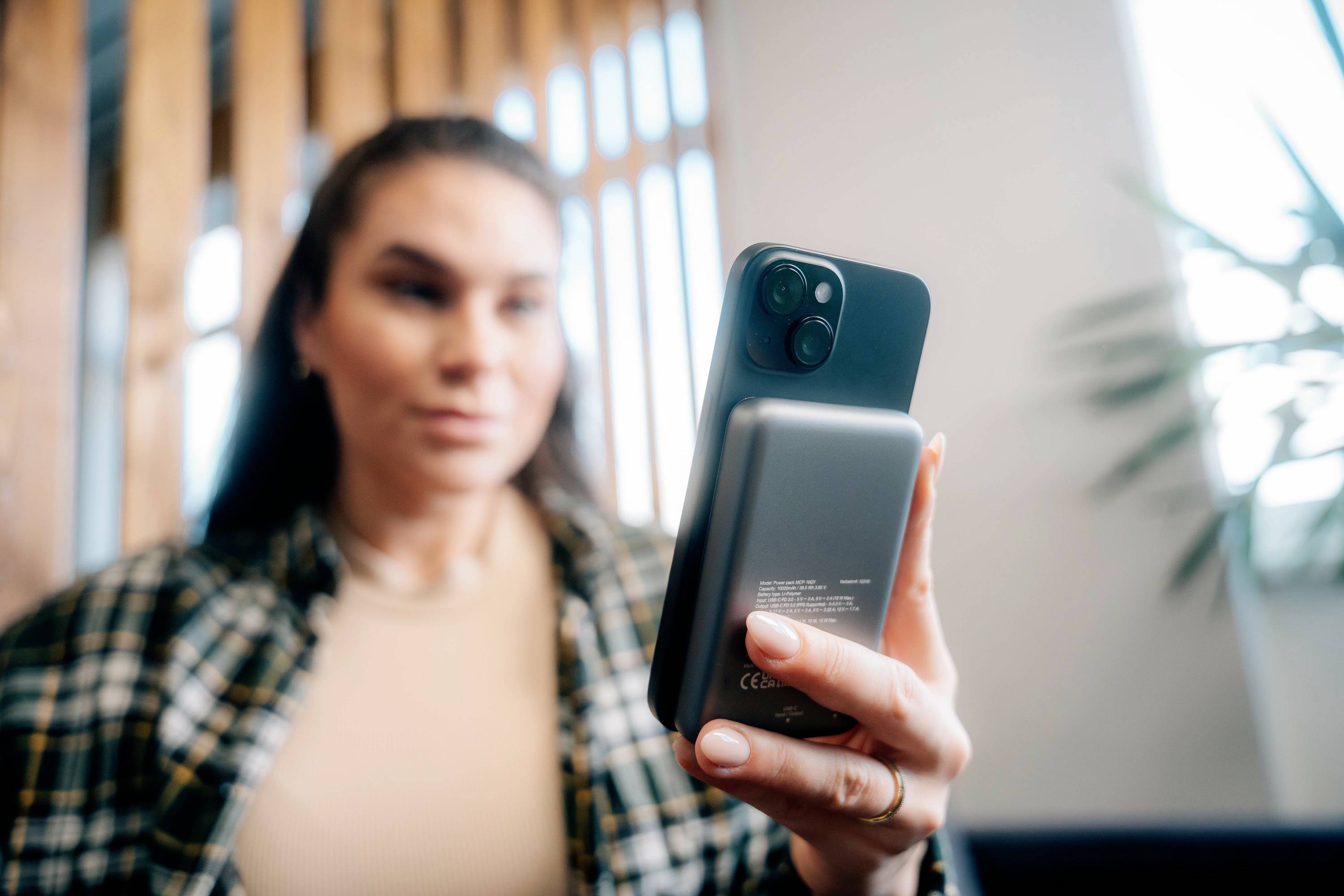 Eine Person hält ein Smartphone mit einer angehängten Powerbank, sitzt in einem Café. Die Person ist im Hintergrund unscharf.