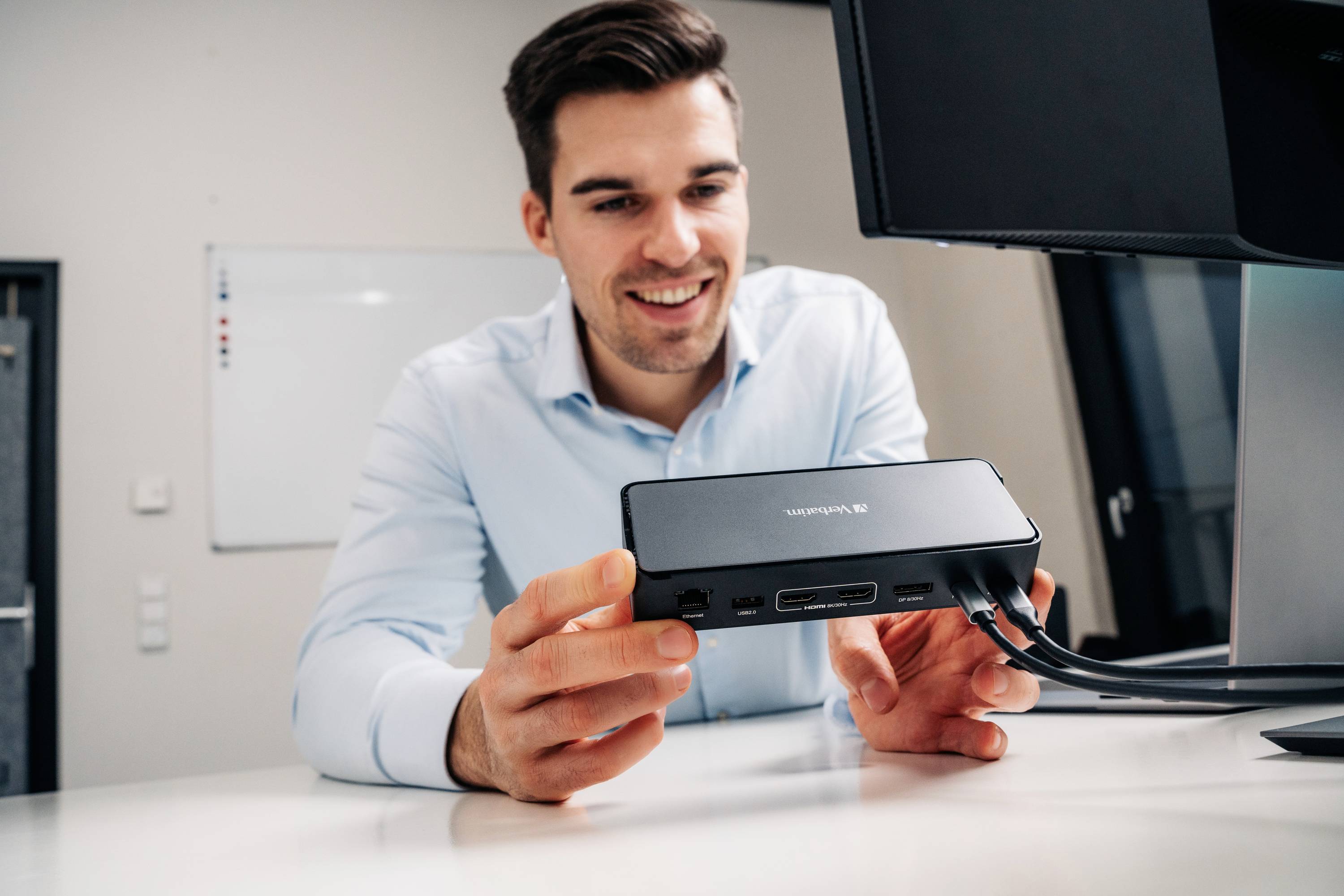 Ein lächelnder Mann sitzt an einem Tisch und hält eine Dockingstation. Im Hintergrund sind ein Whiteboard und ein Monitor sichtbar.