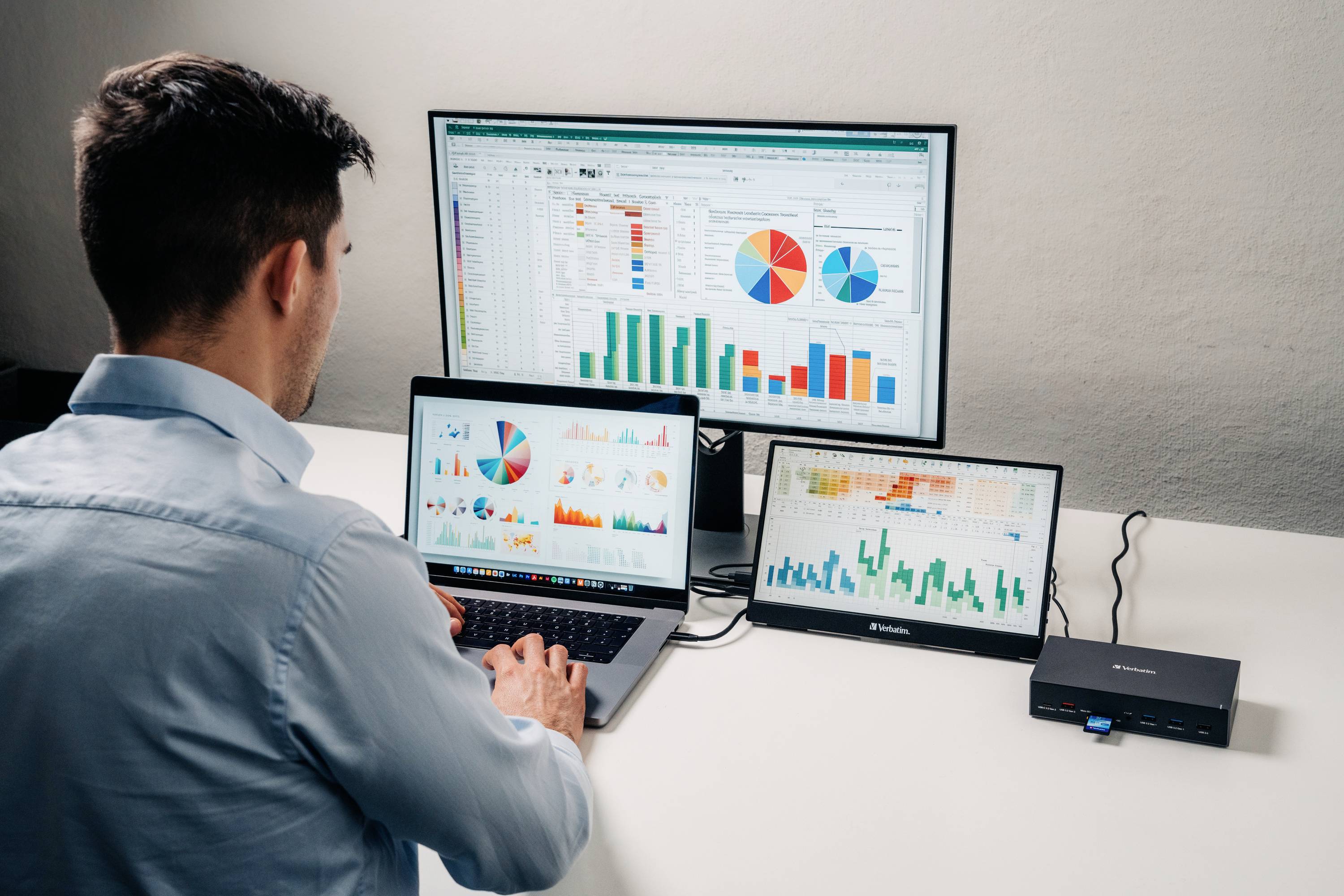 Ein Mann arbeitet an einem Schreibtisch mit einem Laptop und zwei Monitoren, die Diagramme und Datenanalysen anzeigen.