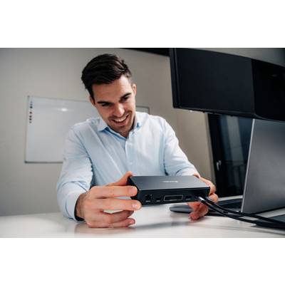 Ein Mann in einem hellblauen Hemd sitzt an einem Schreibtisch und hält lächelnd ein elektronisches Gerät mit mehreren Anschlüssen. Im Hintergrund sind ein Monitor und ein Whiteboard zu sehen.