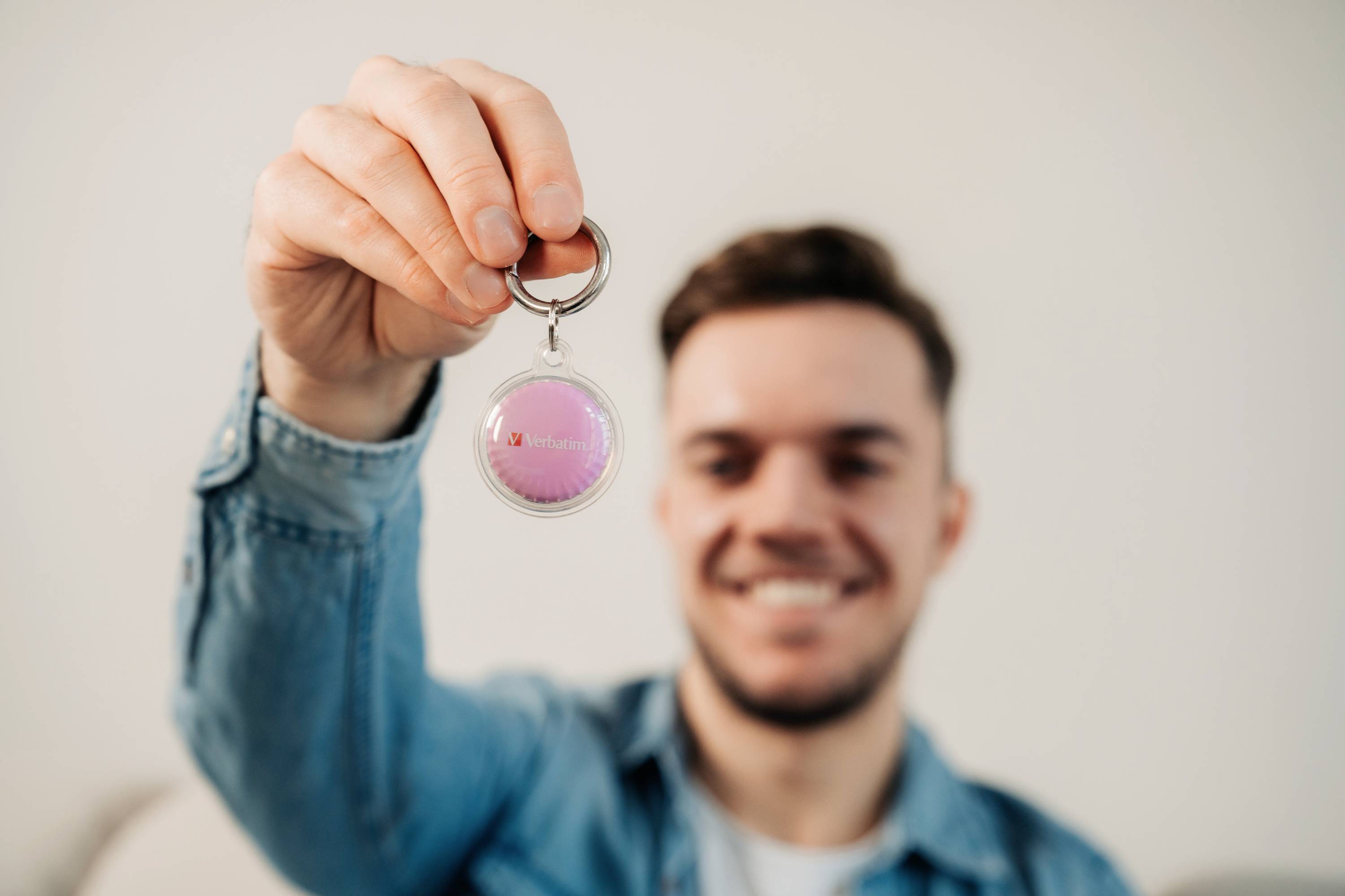 Ein Mann hält einen Schlüsselanhänger mit einer pinken Fläche und einem kleinen roten Herzsymbol in die Kamera. Hintergrund unscharf.