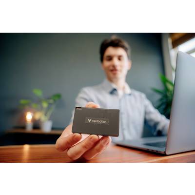 Eine Person am Schreibtisch hält eine externe Festplatte von Verbatim in die Kamera, während sie an einem Laptop arbeitet.