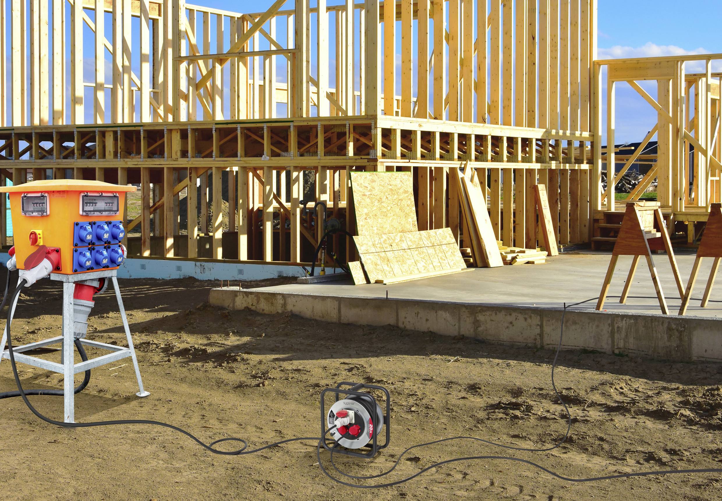 Eine Baustelle mit Holzrahmenarbeiten in Gange. Eine Elektroverteilerdose und eine Kabeltrommel befinden sich im Vordergrund und versorgen die Baustelle mit Strom.