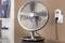 Ein Tischventilator mit Metallgitter steht auf einem Tisch. Rechts daneben ist ein an der Wand montierter Steckdosen-Timer sichtbar.