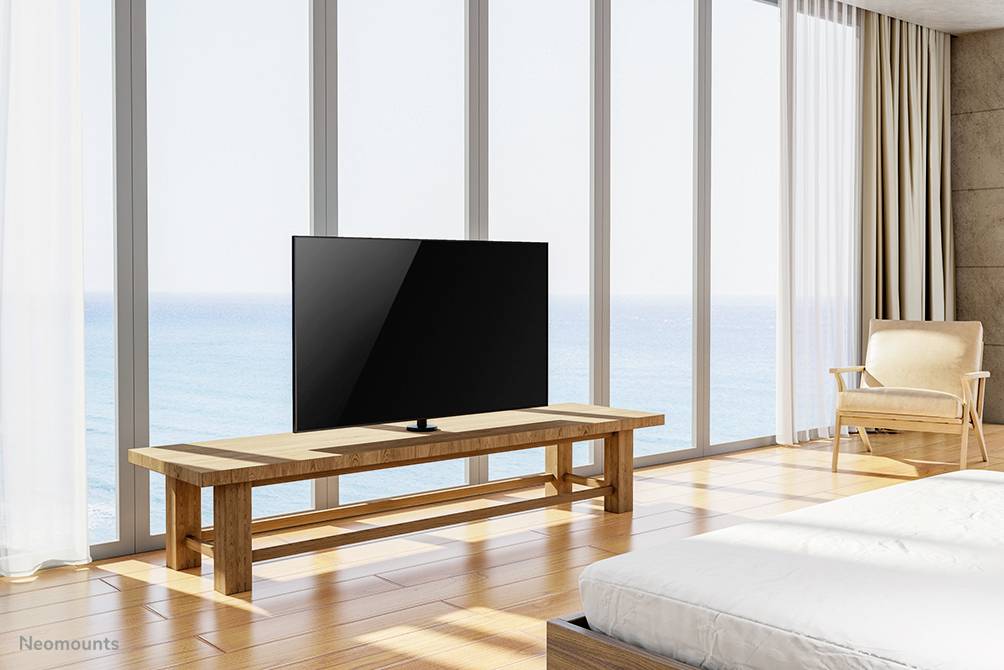 Ein moderner Raum mit großen Fenstern zeigt einen Fernseher auf einem Holztisch, mit Blick auf das Meer. Ein Sessel steht daneben.