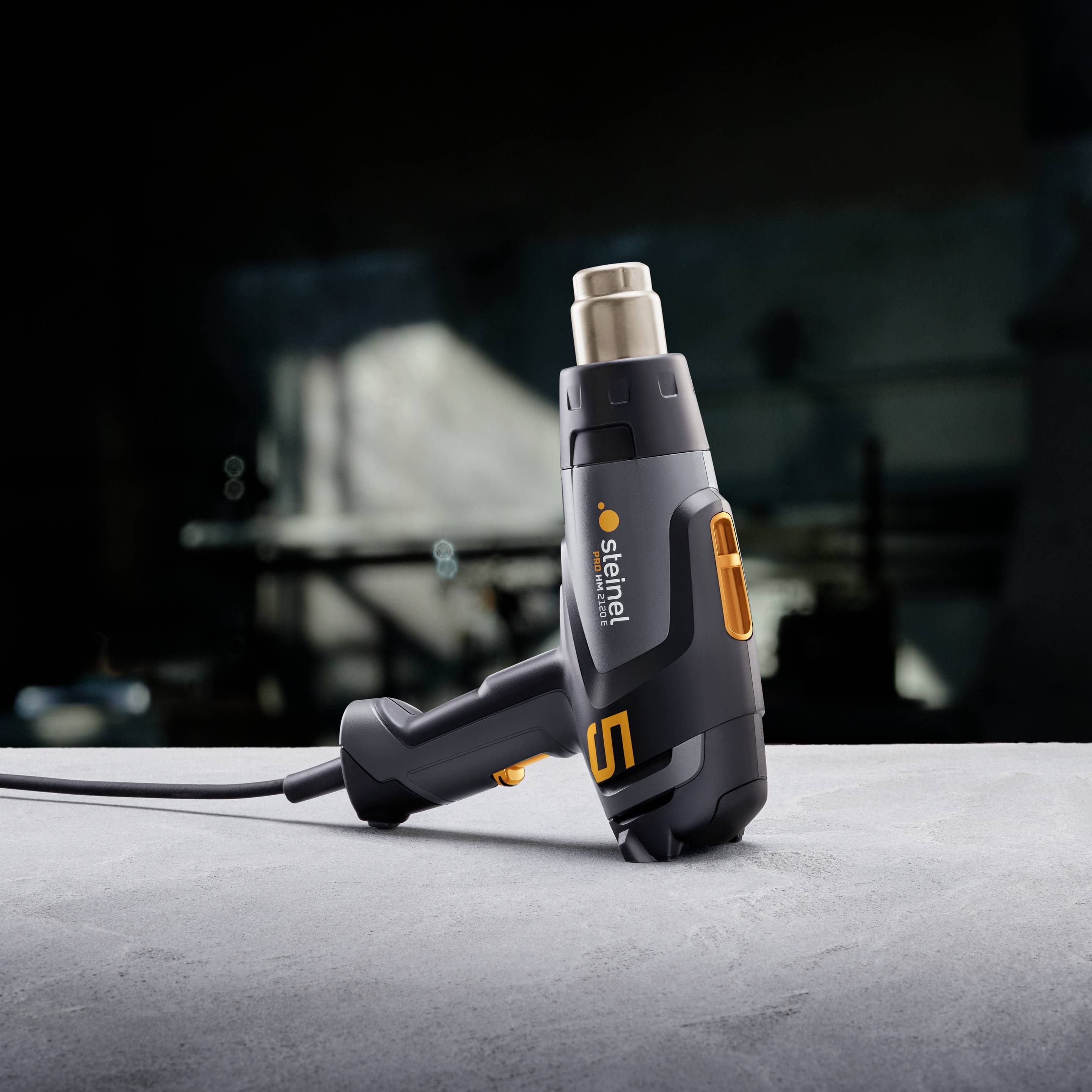 Heißluftgebläse auf einem Tisch, im Vordergrund. Das Gerät ist schwarz-grau mit einem orangenen Akzent und zeigt die Marke 'Steinel'.