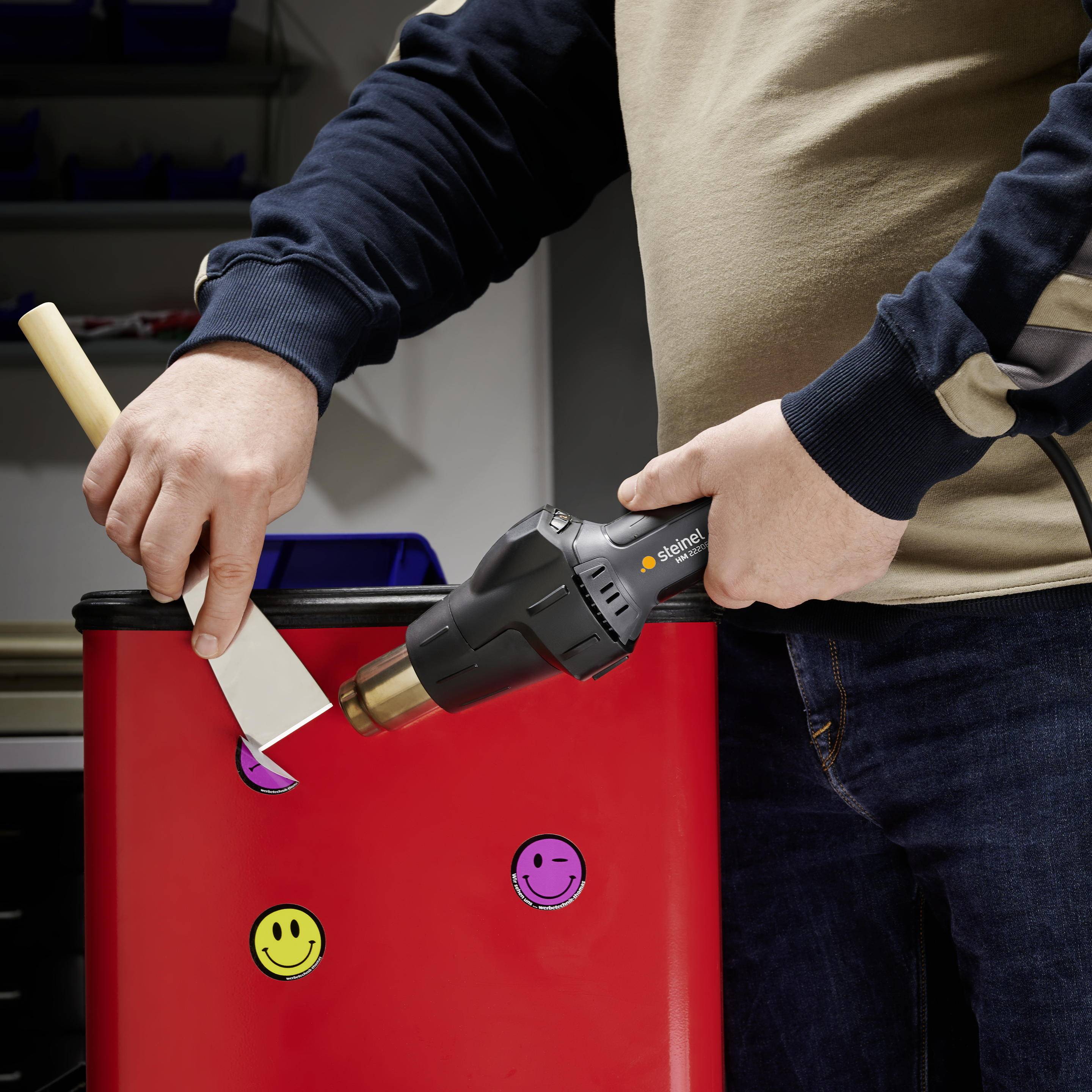 Person streicht rote Farbe auf Kantholz mit zwei gelben Smileys. Hintergrund zeigt Regale.