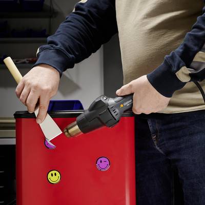Person streicht rote Farbe auf Kantholz mit zwei gelben Smileys. Hintergrund zeigt Regale.