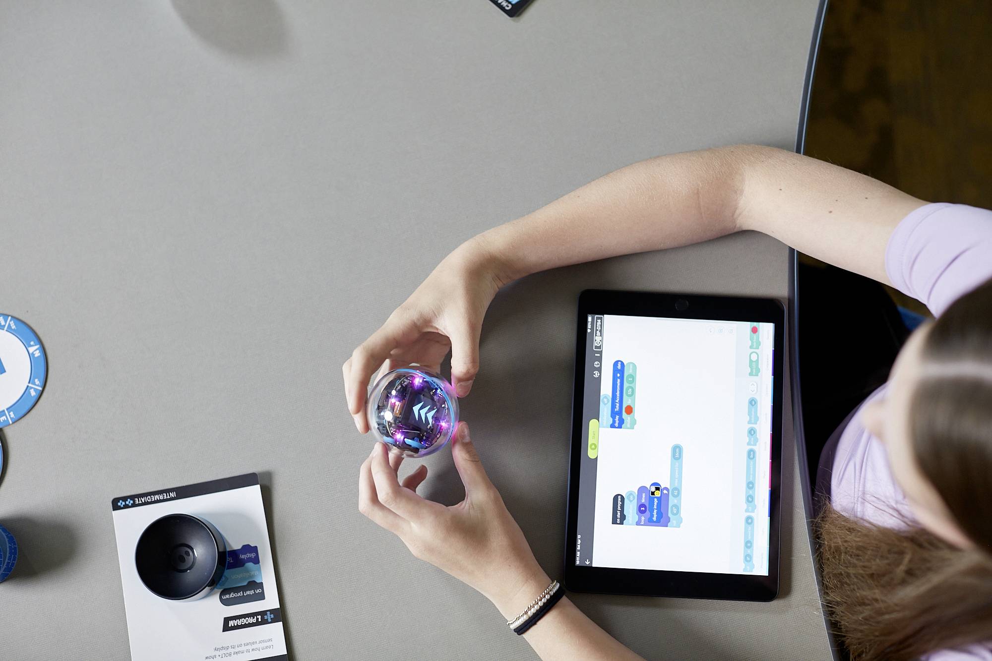 Person hält einen programmierbaren Ball und nutzt ein Tablet mit einer App zum Codieren. Flyer und weiterer Ball liegen auf dem Tisch.