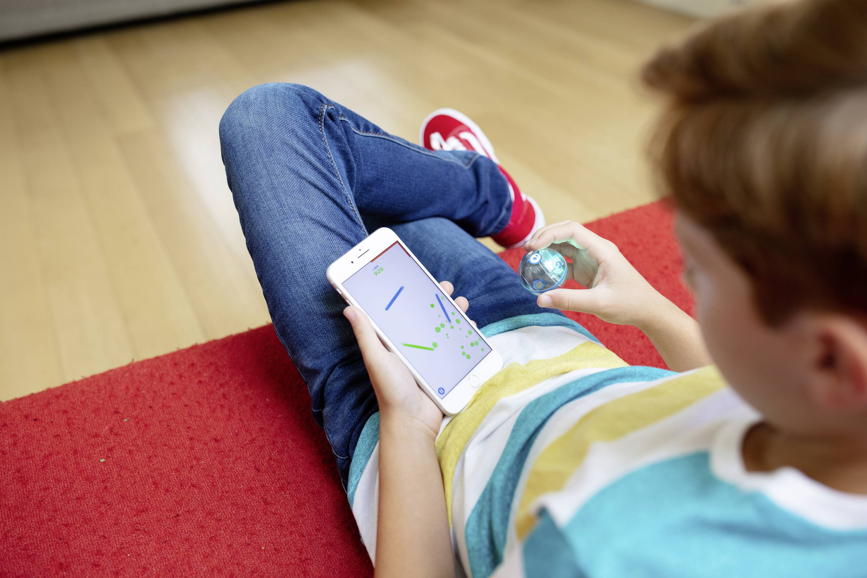 Ein Kind spielt ein mobiles Spiel auf einem Smartphone, während es einen kleinen blauen Würfel hält. Es sitzt auf einem roten Teppich.