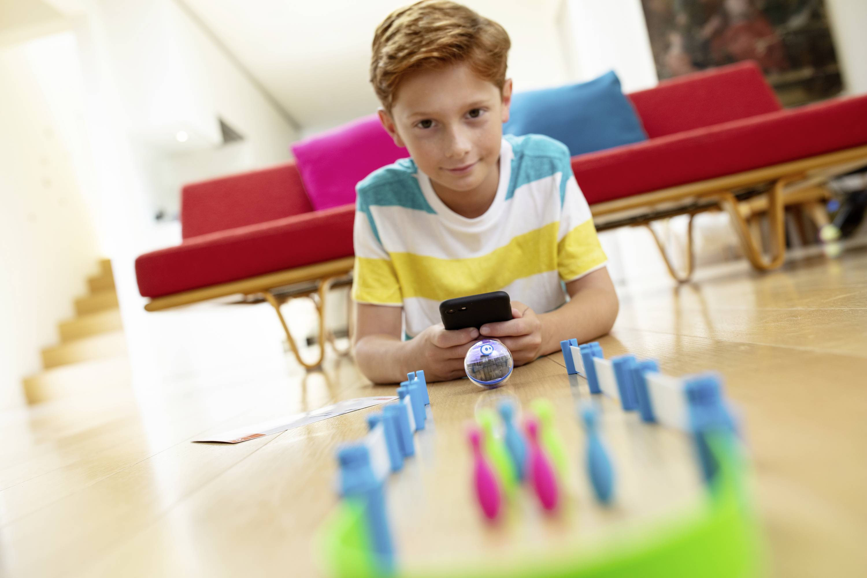 Ein Junge liegt auf dem Boden und steuert ein Spielzeug-Roboterball mit einem Smartphone. Im Hintergrund sind bunte Kissen auf einem Sofa.