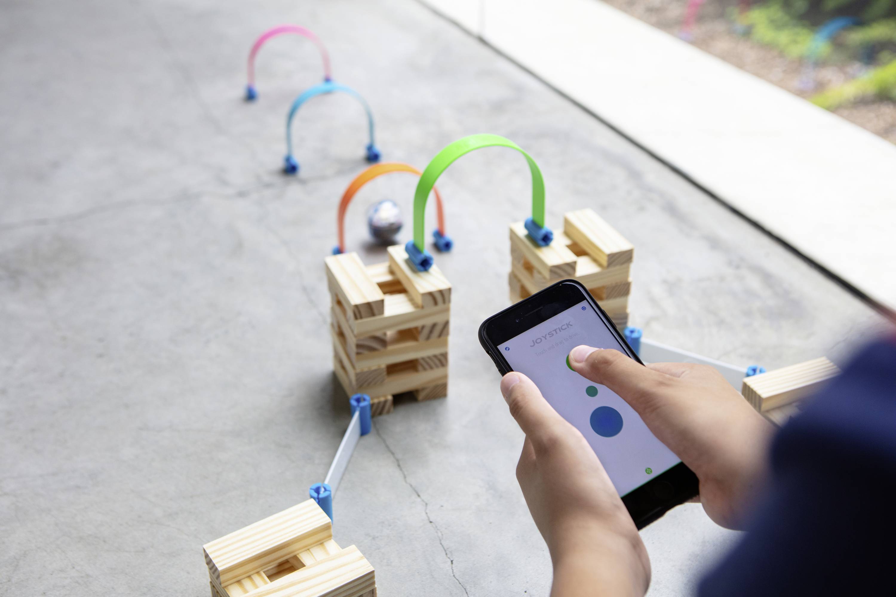Eine Person steuert mithilfe eines Smartphones einen kleinen Roboter durch einen Parcours aus Holzblöcken und bunten Bögen.
