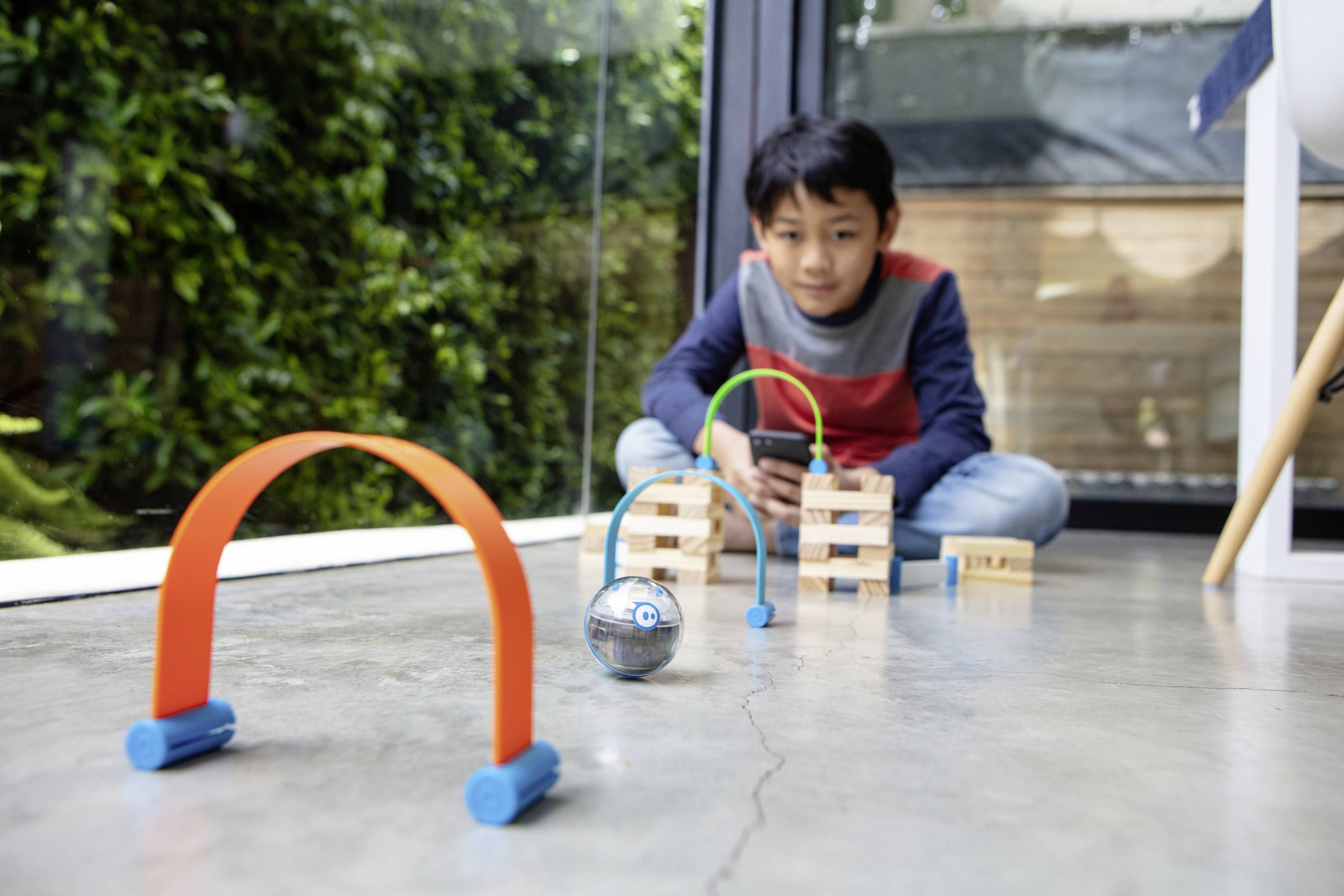 Ein Junge spielt auf dem Boden mit einem Roboterball, der durch bunte Tore rollt. Im Hintergrund sind Glasfenster und Grünpflanzen.