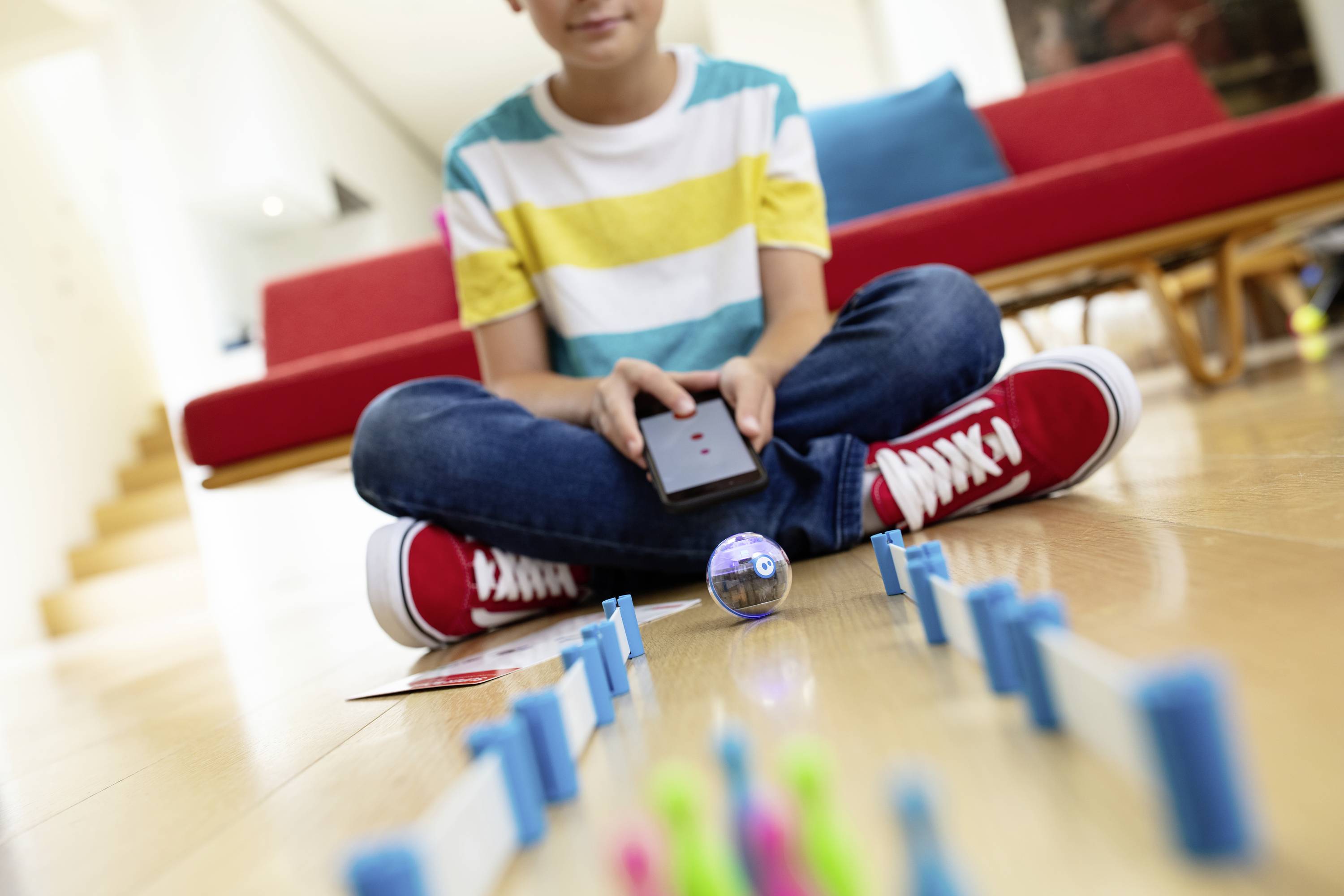 Ein Kind steuert mit einem Smartphone einen kleinen Spielzeugroboter bei einem Hindernisparcours. Im Hintergrund ein rotes Sofa.