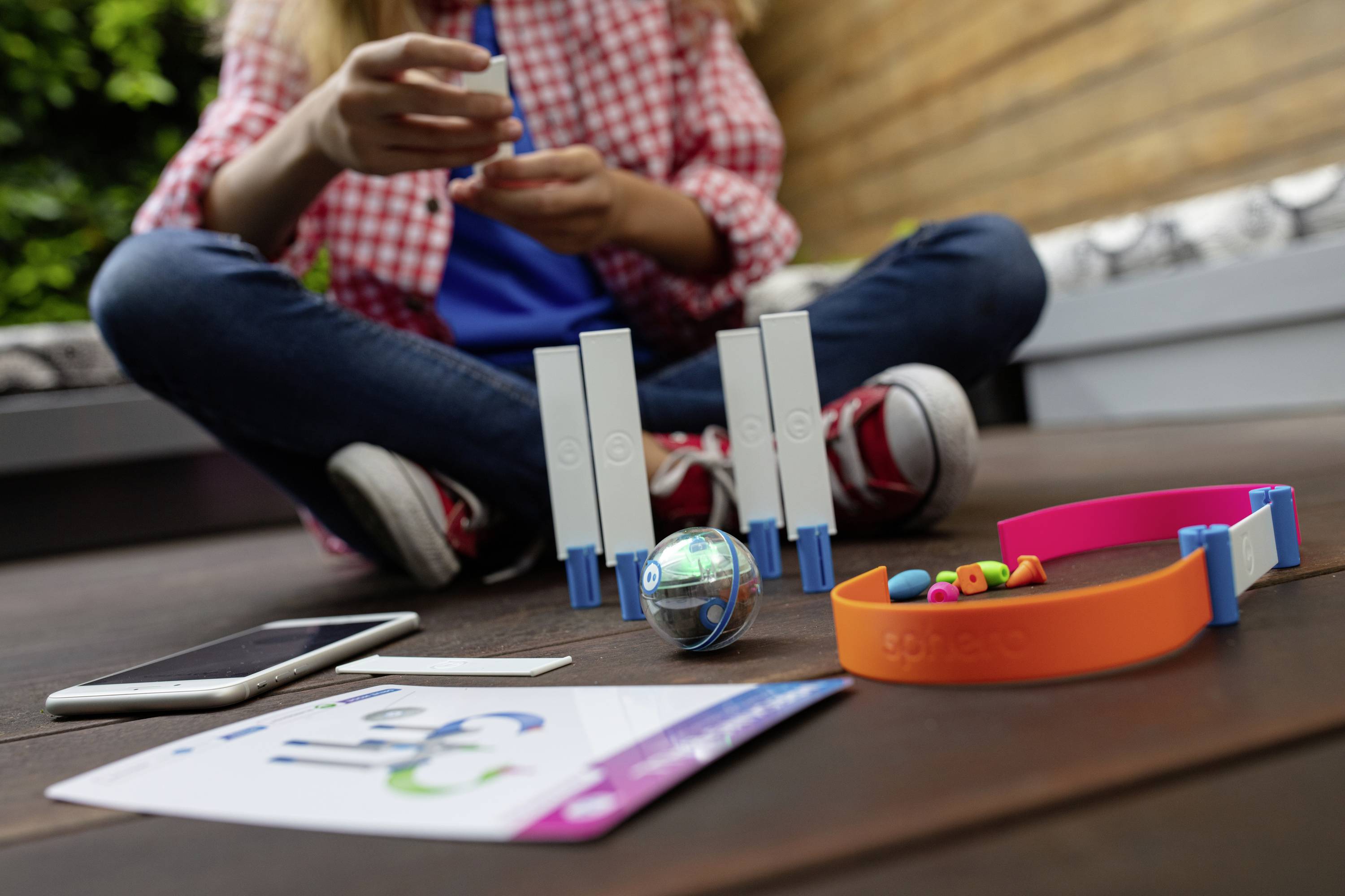 Eine Person sitzt im Schneidersitz auf einem Boden. Vor ihr liegen ein Smartphone, bunte Armbänder und ein kleiner, transparenter Roboterball.