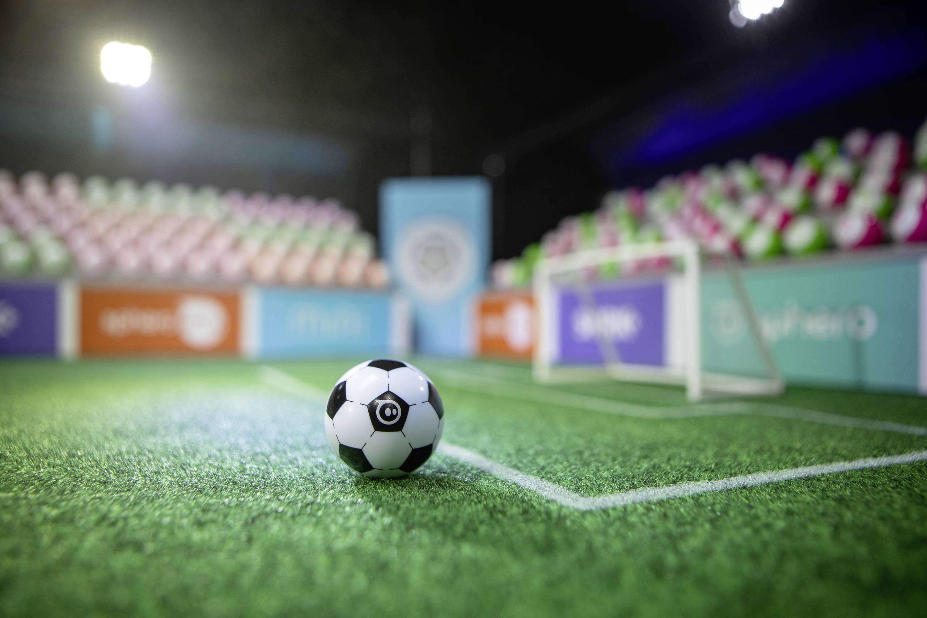 Ein schwarzer-weißer Fußball liegt auf einem beleuchteten Kunstrasenplatz, umgeben von bunten Tribünen und kleinen Toren im Hintergrund.