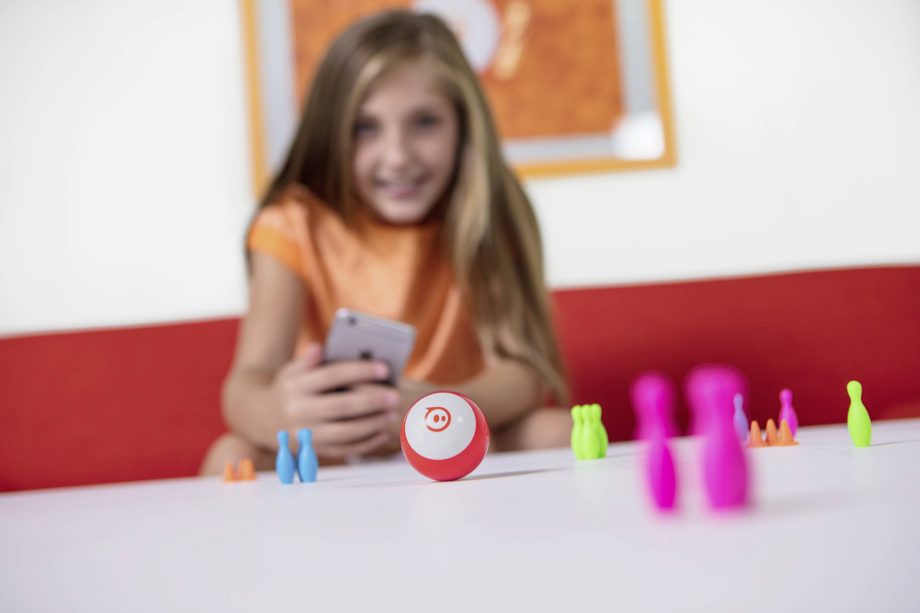 Kind zeigt Freude, spielt mit buntem Spielzeugball auf Tisch; verwendet Smartphone zu Steuerung. Roter Ball im Fokus. 