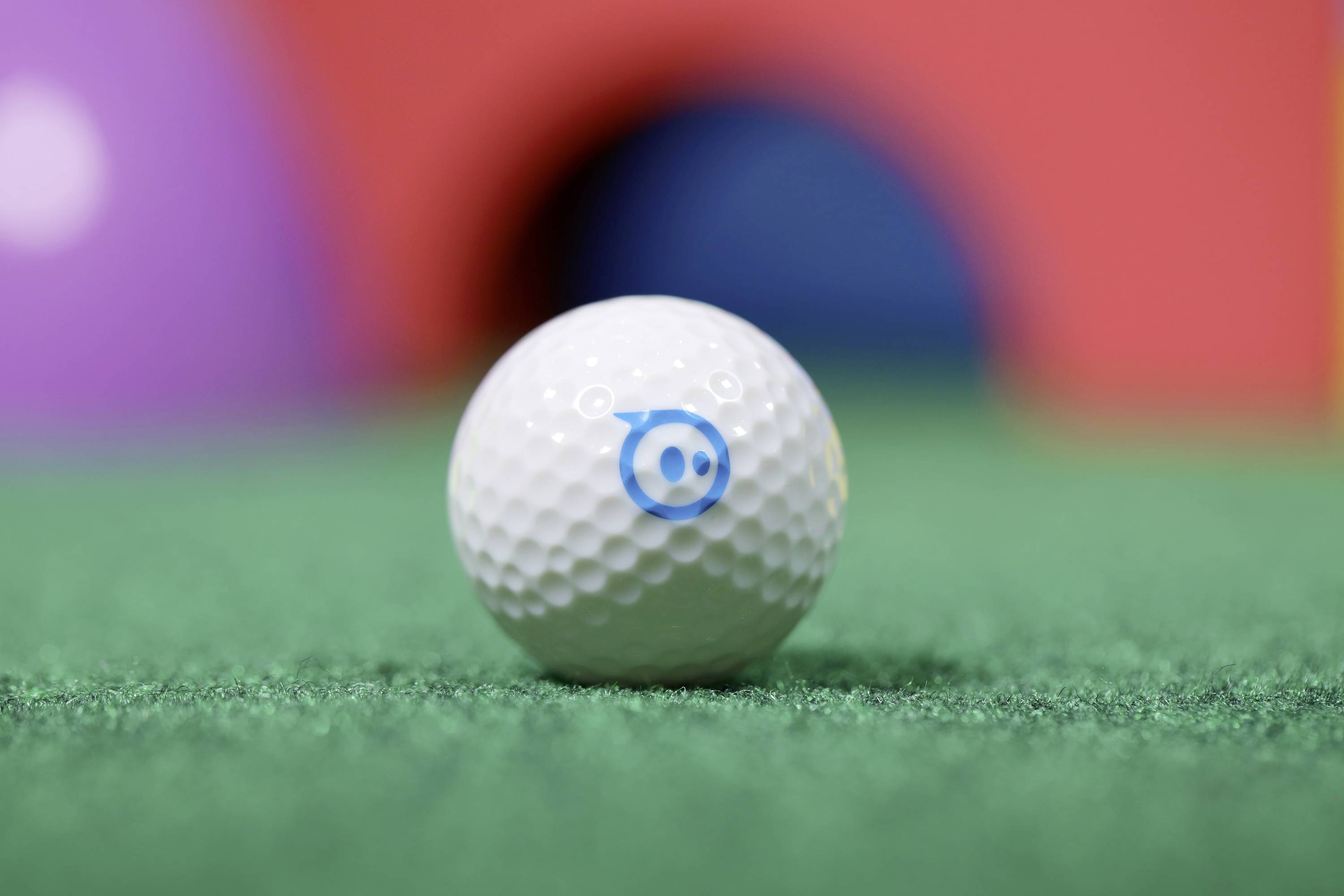 Golfball mit blauem Symbol auf grünem Rasen, verschwommene bunte Hintergründe.
