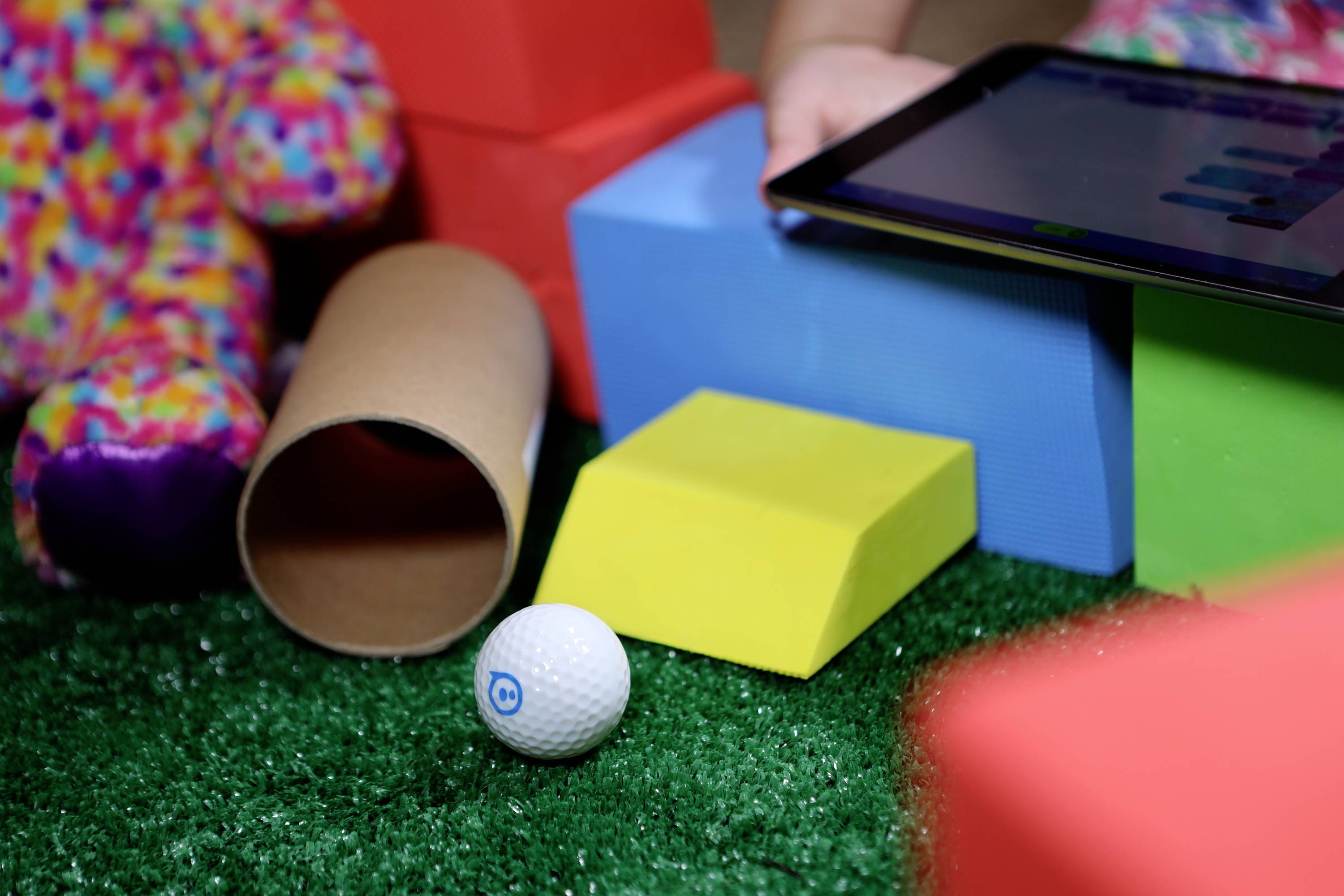 Ein Golfball liegt auf grünem Filz zwischen bunten Bauklötzen und einer Papprolle; daneben ein Tablet mit einem nicht erkennbaren Diagramm.