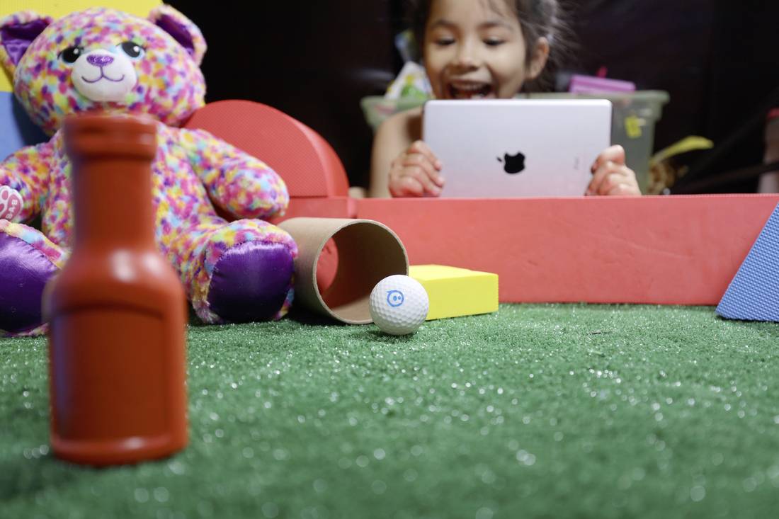 Ein Kind spielt fröhlich mit einem Tablet auf einem Teppich mit Spielzeug, darunter ein bunter Teddybär, ein Plastikfläschchen und ein Golfball.
