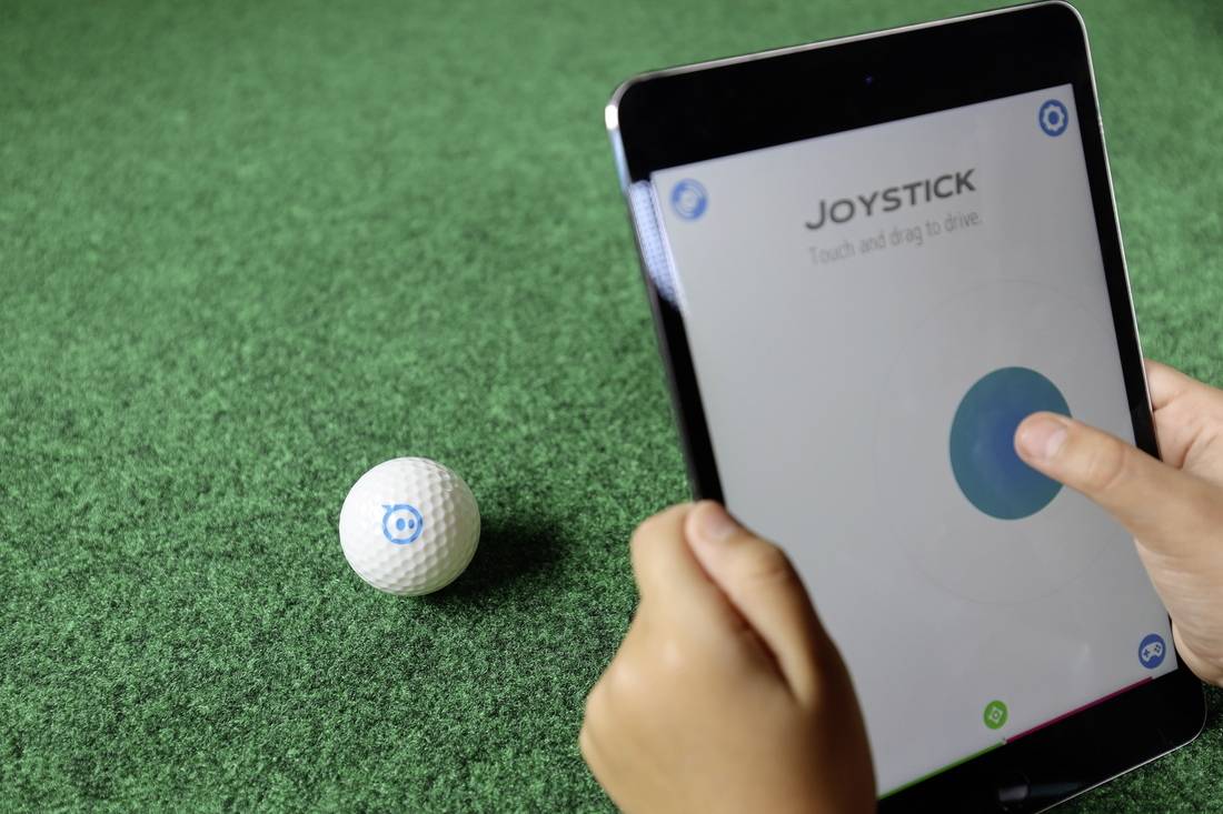 Person steuert mit einem Tablet einen kleinen, kugelförmigen Roboter auf grünem Teppich. Auf dem Bildschirm steht 'JOYSTICK'.
