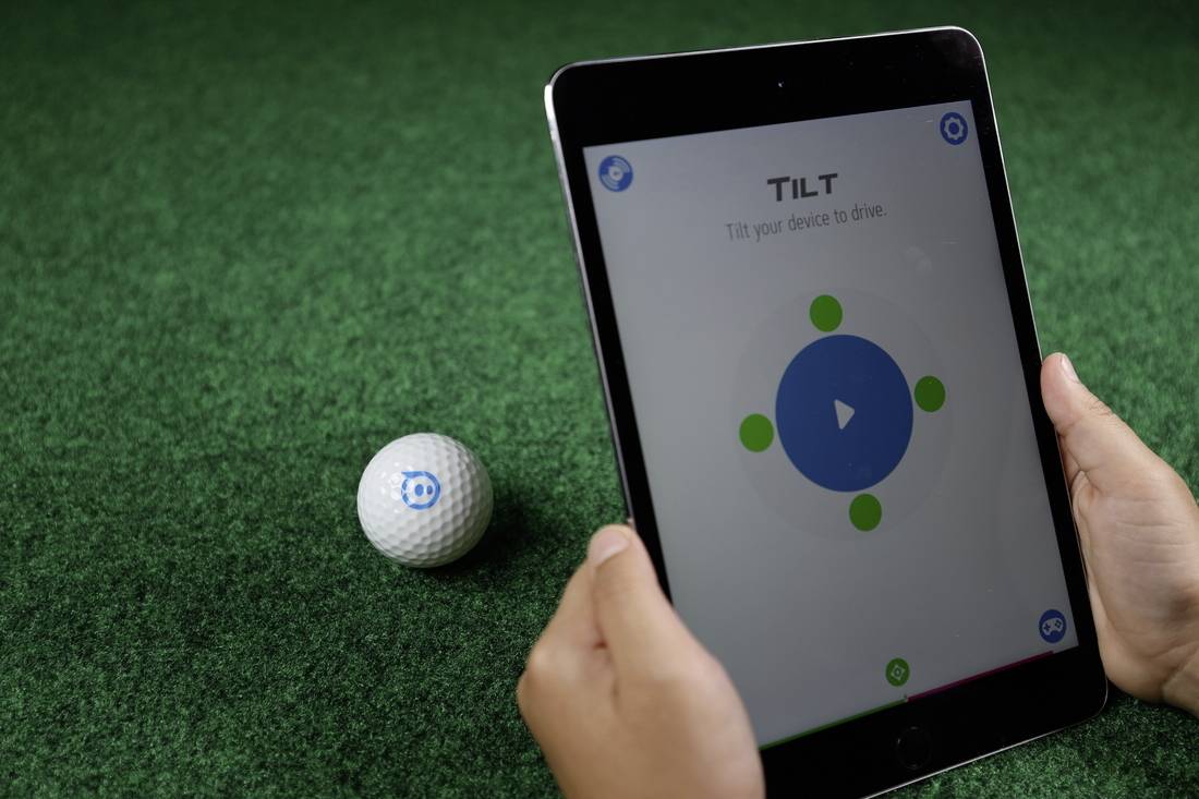 Ein Tablet zeigt eine Steuerungs-App mit Anweisungen. Ein Golfball mit einem Logo liegt daneben auf grünem Kunstrasen.