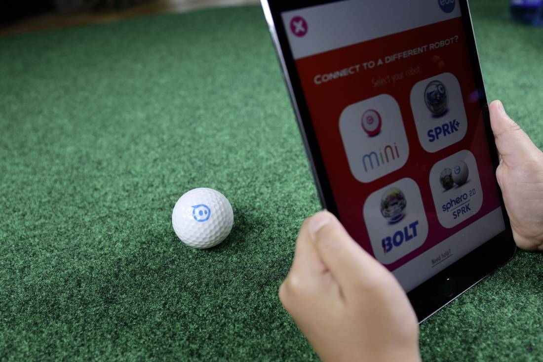 Eine Person hält ein Tablet, das eine App zur Verbindung mit einem Roboter zeigt. Vor dem Tablet liegt ein kleiner, ballförmiger Roboter auf grünem Kunstrasen.