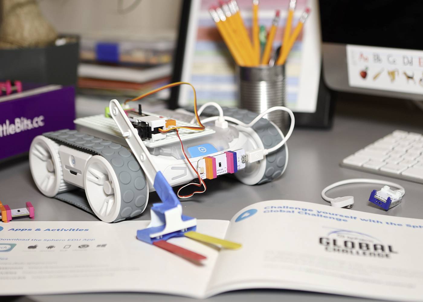 Roboterauto auf Schreibtisch, umgeben von Elektronikkomponenten und Büroartikeln. Es steht neben geöffnetem Buch für kreative Projekte.