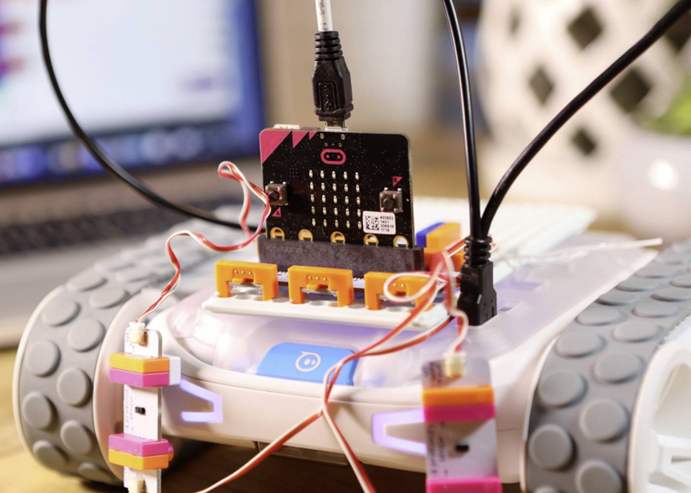 Ein Roboterauto mit einer Mikrocontroller-Platine ist über Kabel und Stecker verbunden. Im Hintergrund ist ein Laptop leicht erkennbar.