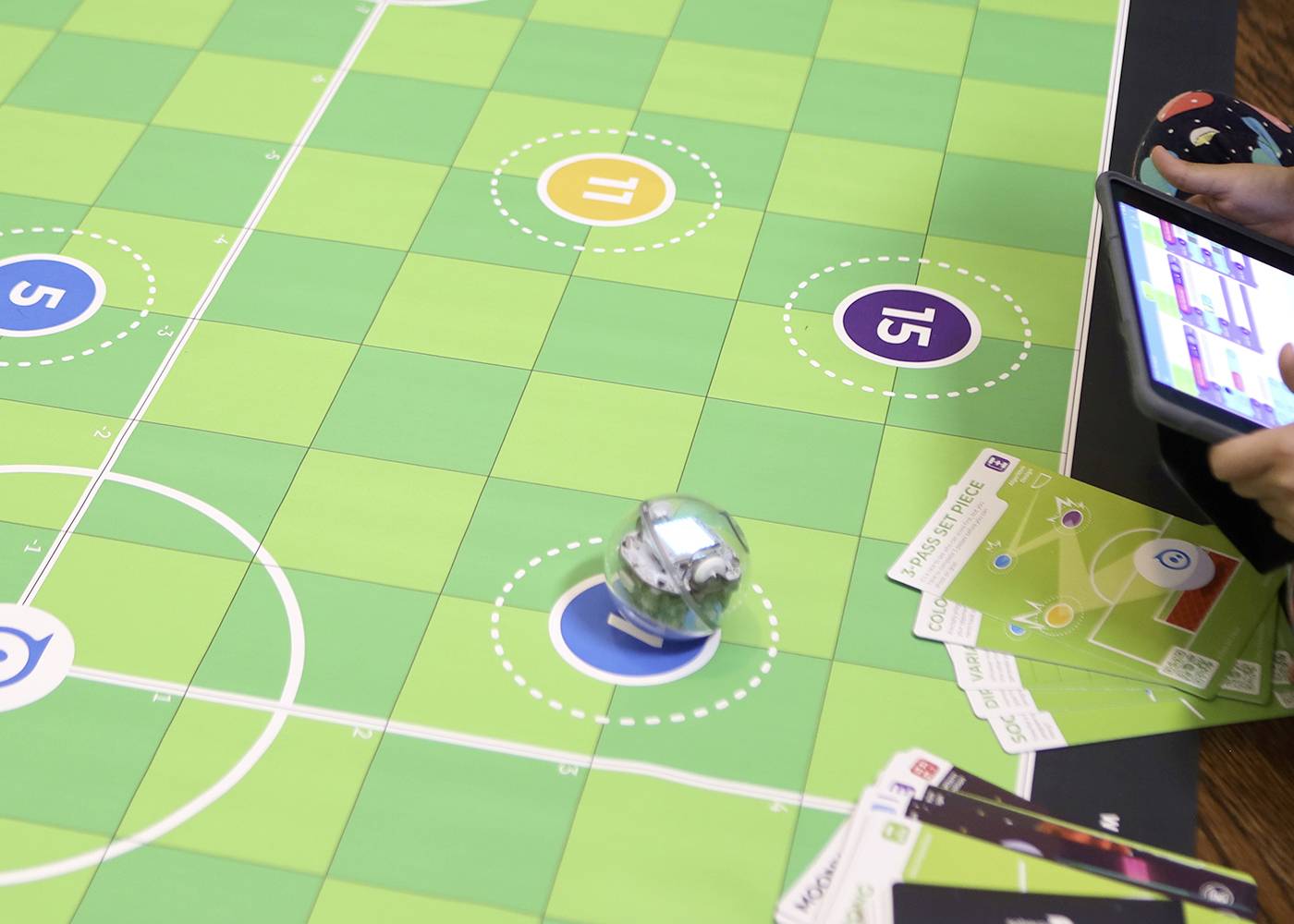 Ein Spielfeld mit nummerierten Kreisen, einem rollenden kleinen Roboter, Karten und einem Tablet. Kinder spielen ein Lernspiel.