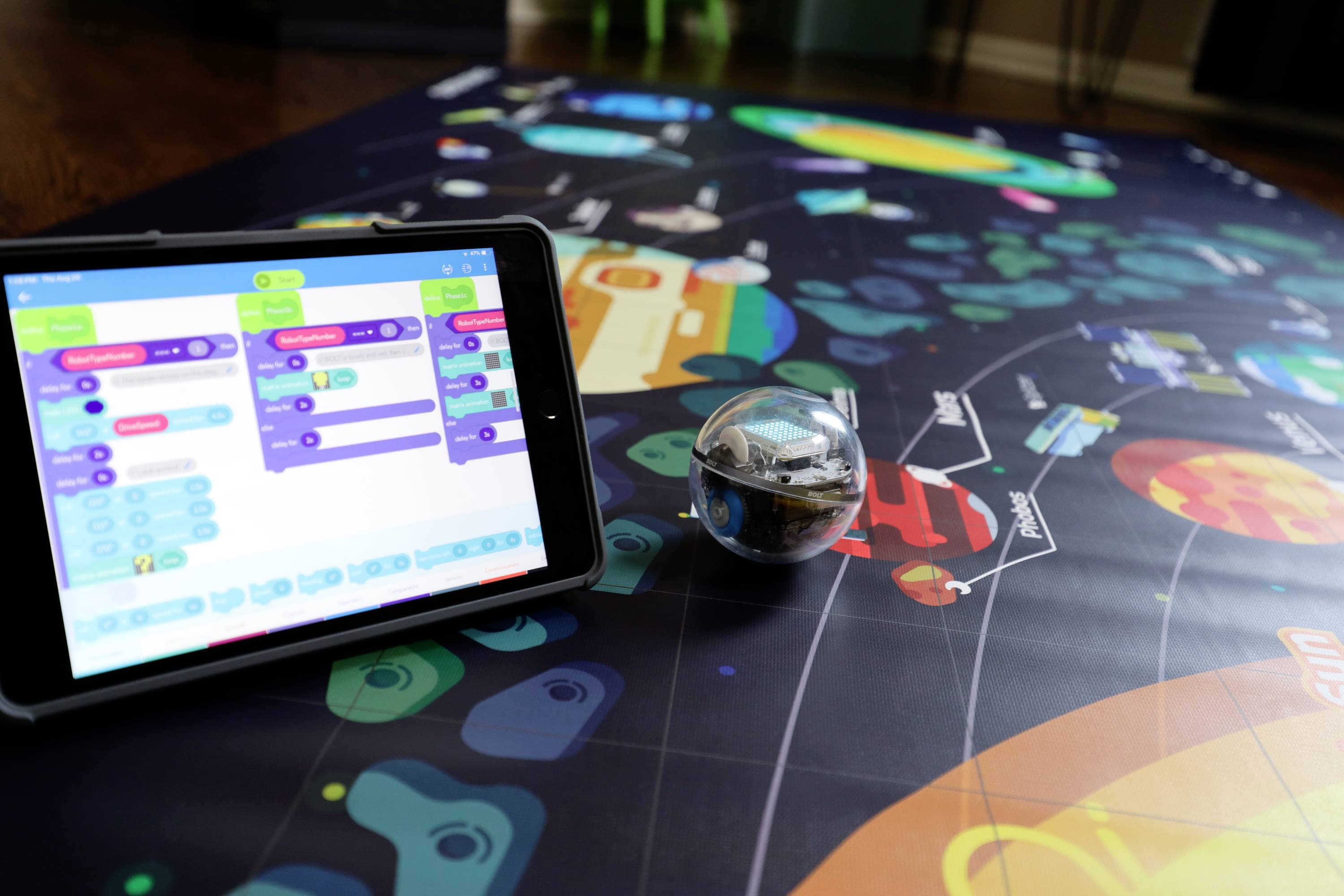 Tablet zeigt Programmier-App neben sphärischem Roboter auf buntem Lernposter, das Planeten und Pfade darstellt.
