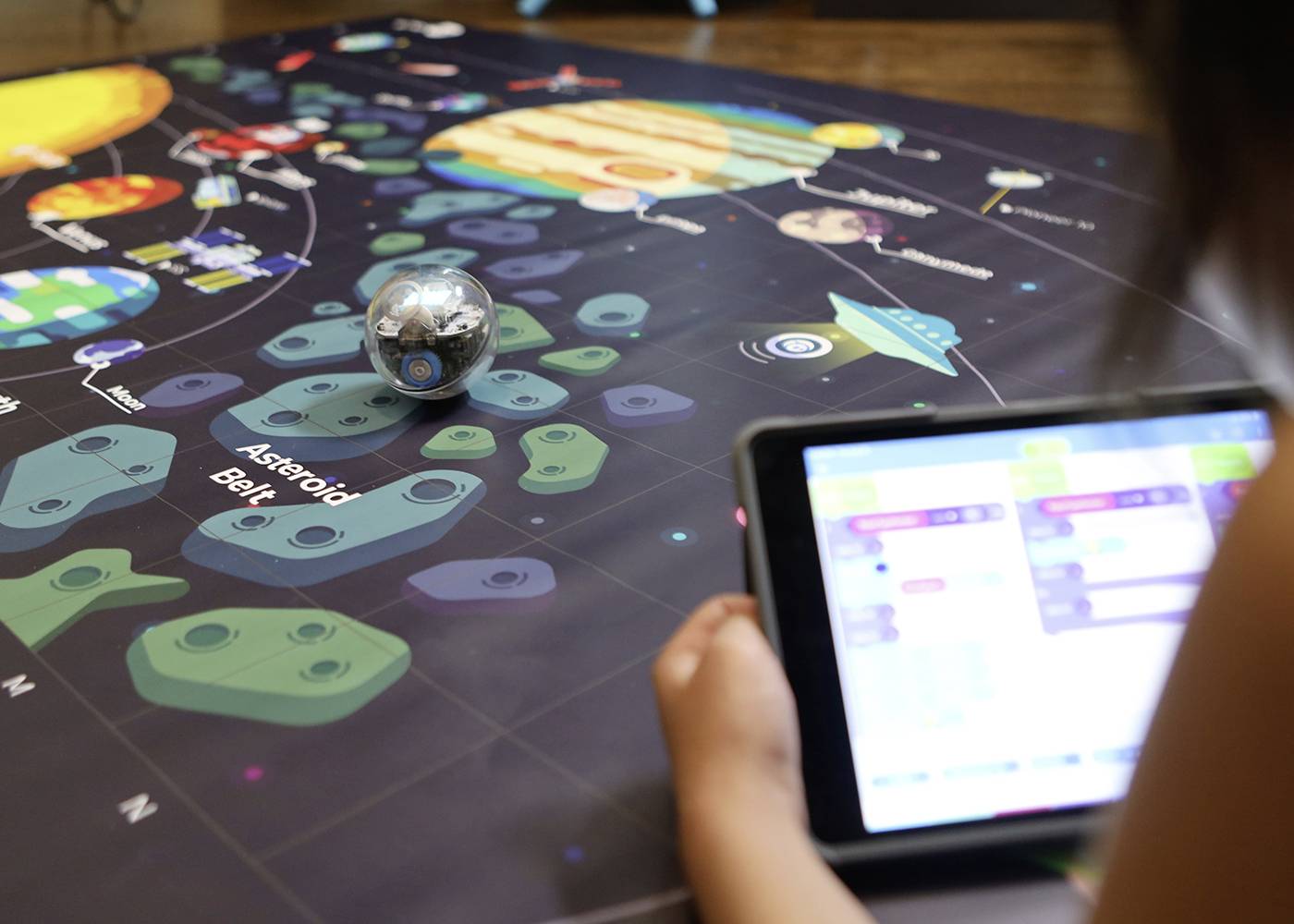 Ein Kind steuert eine Kugel auf einem Weltraumspielbrett mit einem Tablet. Das Brett zeigt Planeten und Asteroidenpfade.