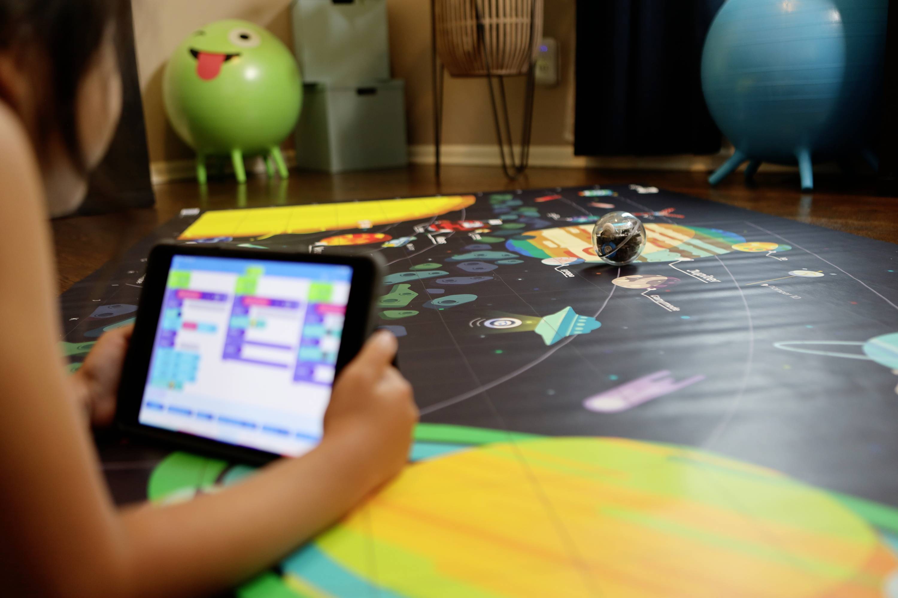 Ein Kind programmiert einen Roboterball mit einem Tablet auf einer bunten Weltraummatte.