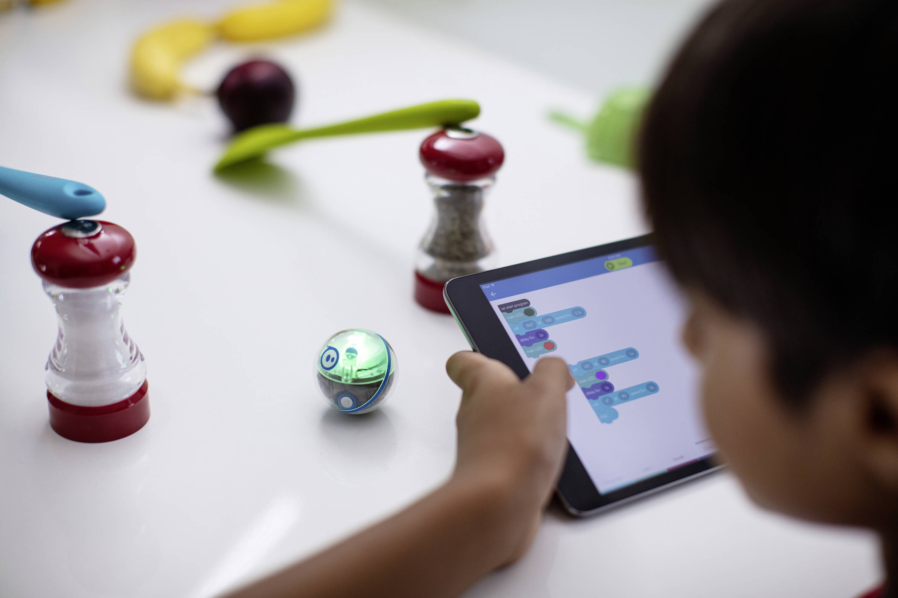 Ein Kind programmiert mit einem Tablet einen kleinen, kugelförmigen Roboter zwischen Gewürzmühlen auf einem Tisch.