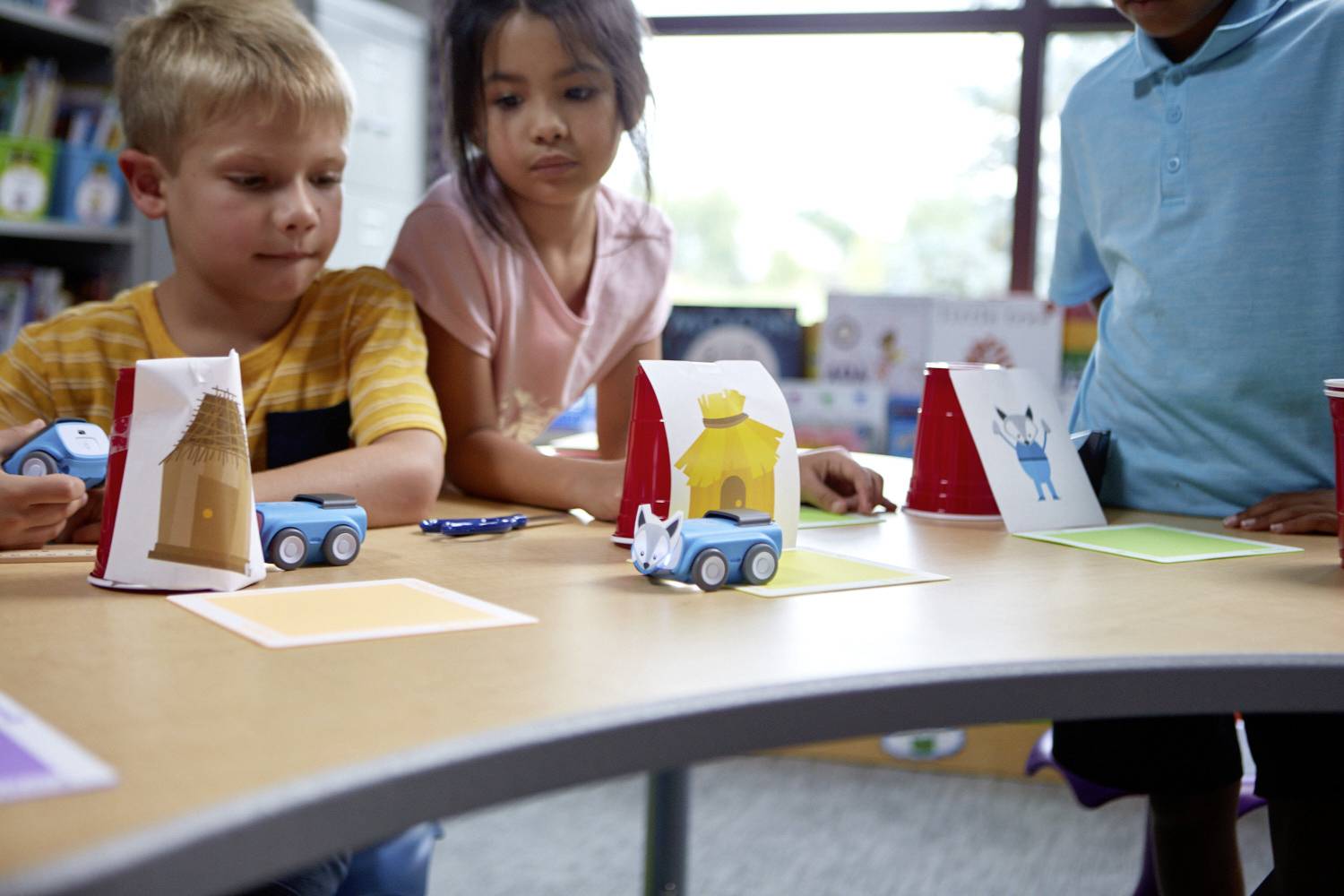 Kinder programmieren kleine Spielzeugautos bei einem Projekt im Unterricht. Die Autos sind auf Karten mit verschiedenen Farben platziert.