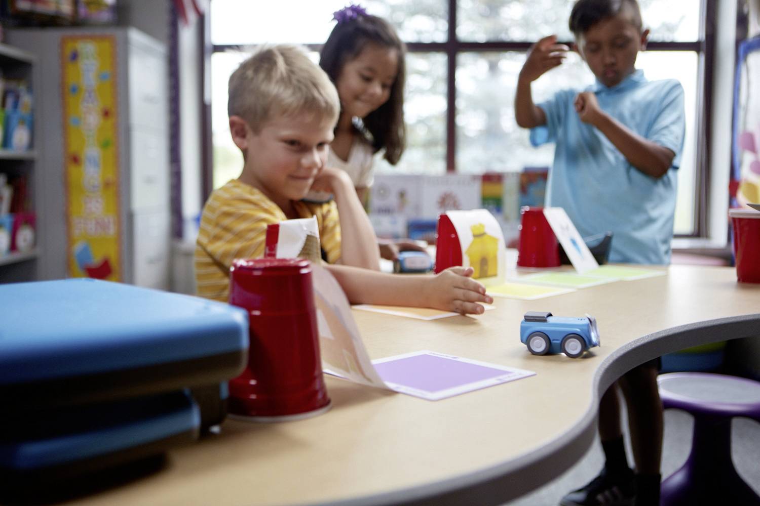 Kinder spielen in einem Klassenzimmer. Ein Kind schiebt ein blaues Spielzeugauto auf einem Tisch. Zwei andere Kinder schauen zu.