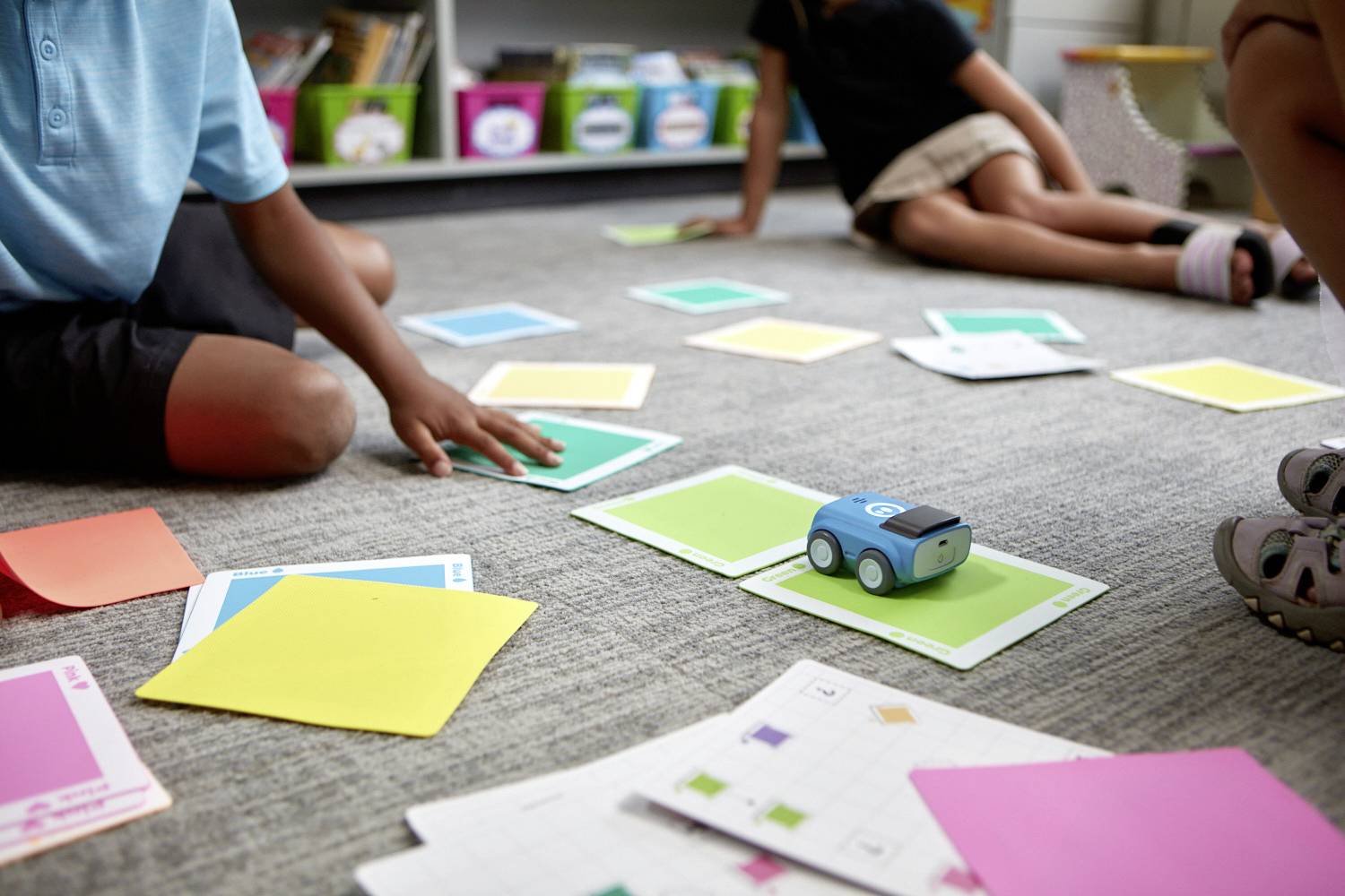 Kinder spielen mit einem kleinen, blauen Spielzeugauto und bunten Karten auf dem Boden in einem Klassenzimmer.